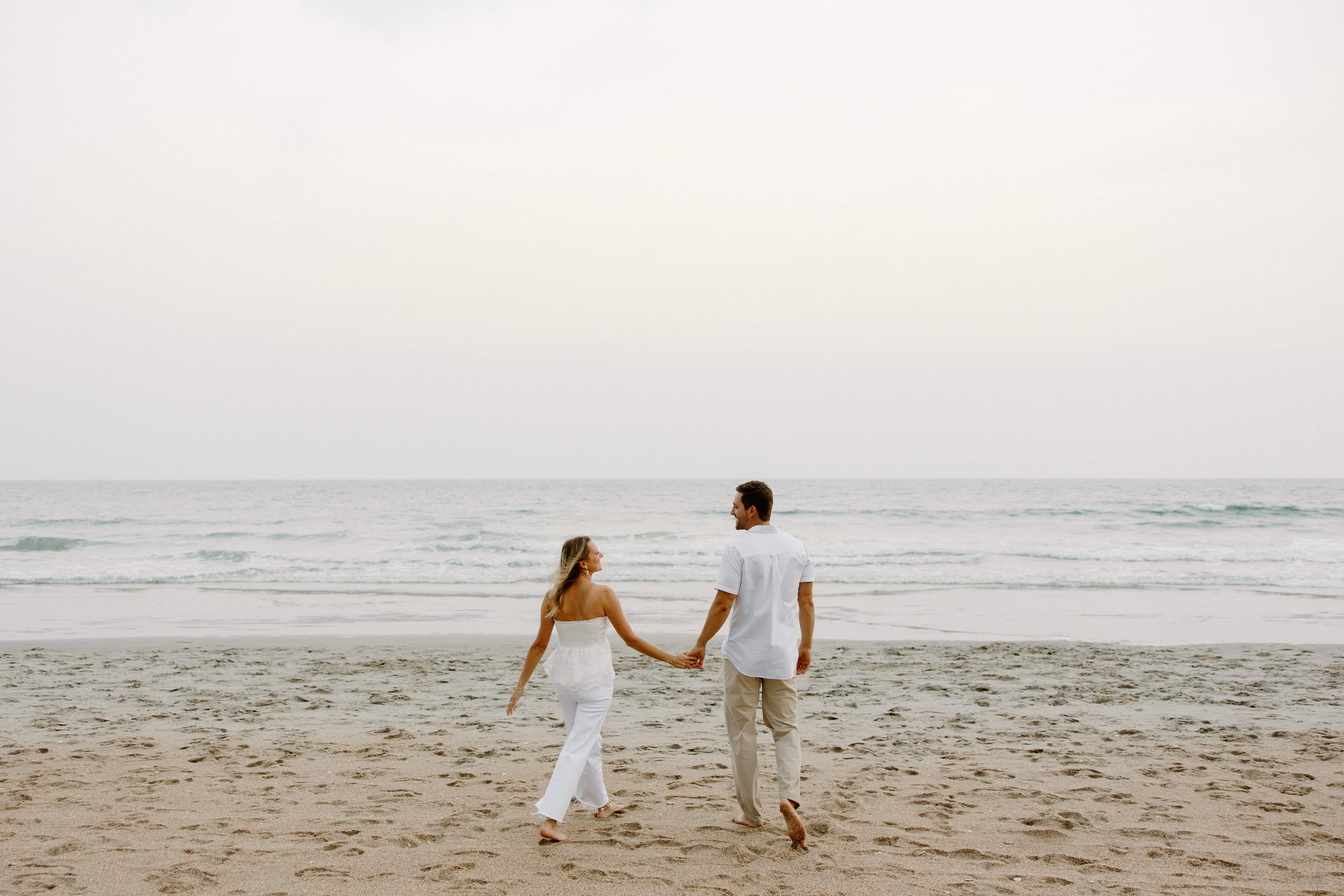  A couple walks away from the camera on a sandy beach, holding hands. The ocean and sky are in the background.