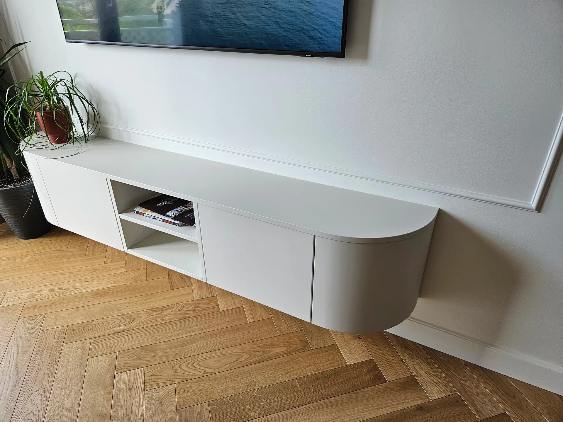 Minimalistyczna szafka pod telewizor z zaokrąglonymi białymi frontami i parkietem w jodełkę.