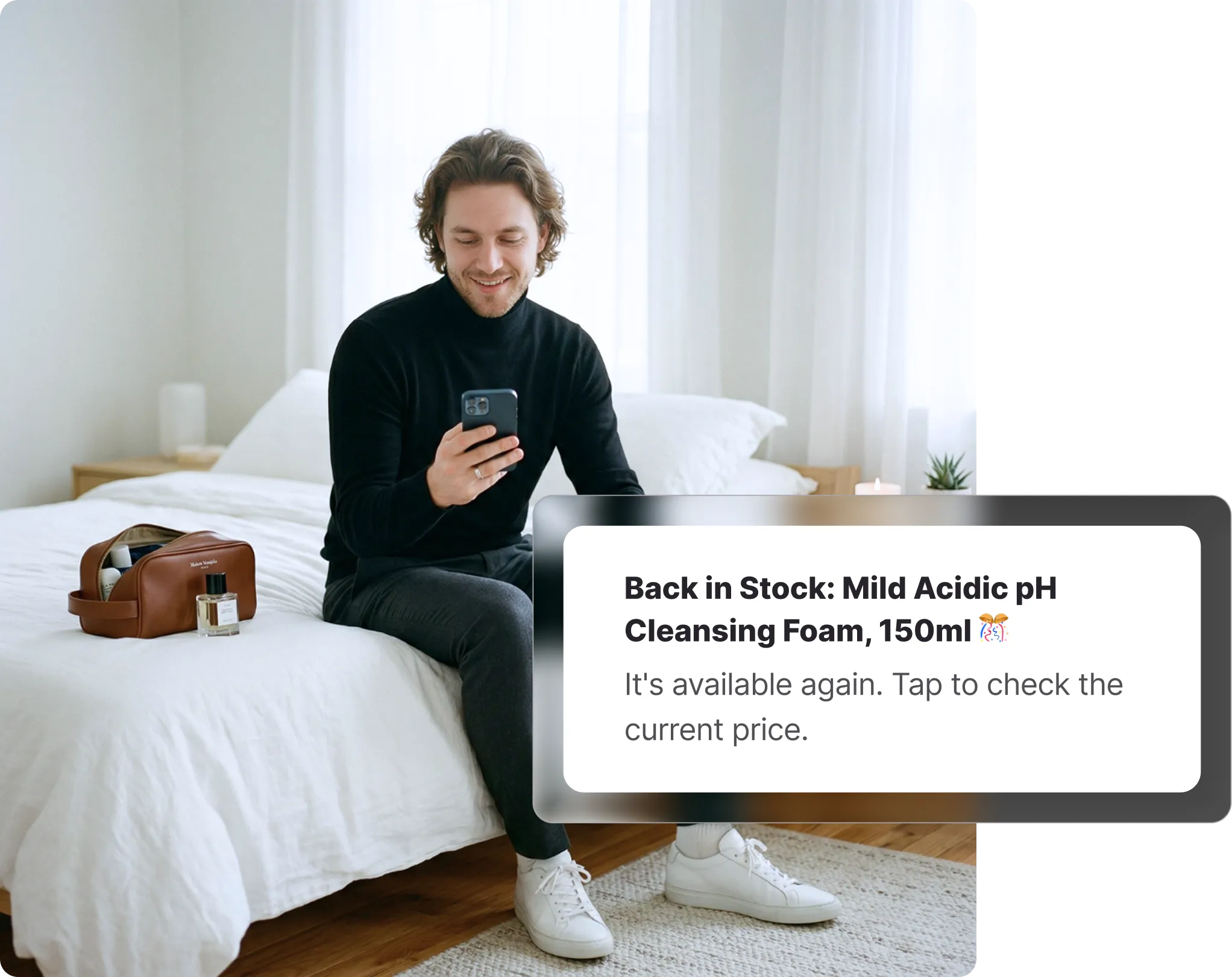 Man sitting on a bed checking his phone with a toiletry bag and fragrance bottle beside him, receiving a YesStyle back-in-stock notification on Whisprice.