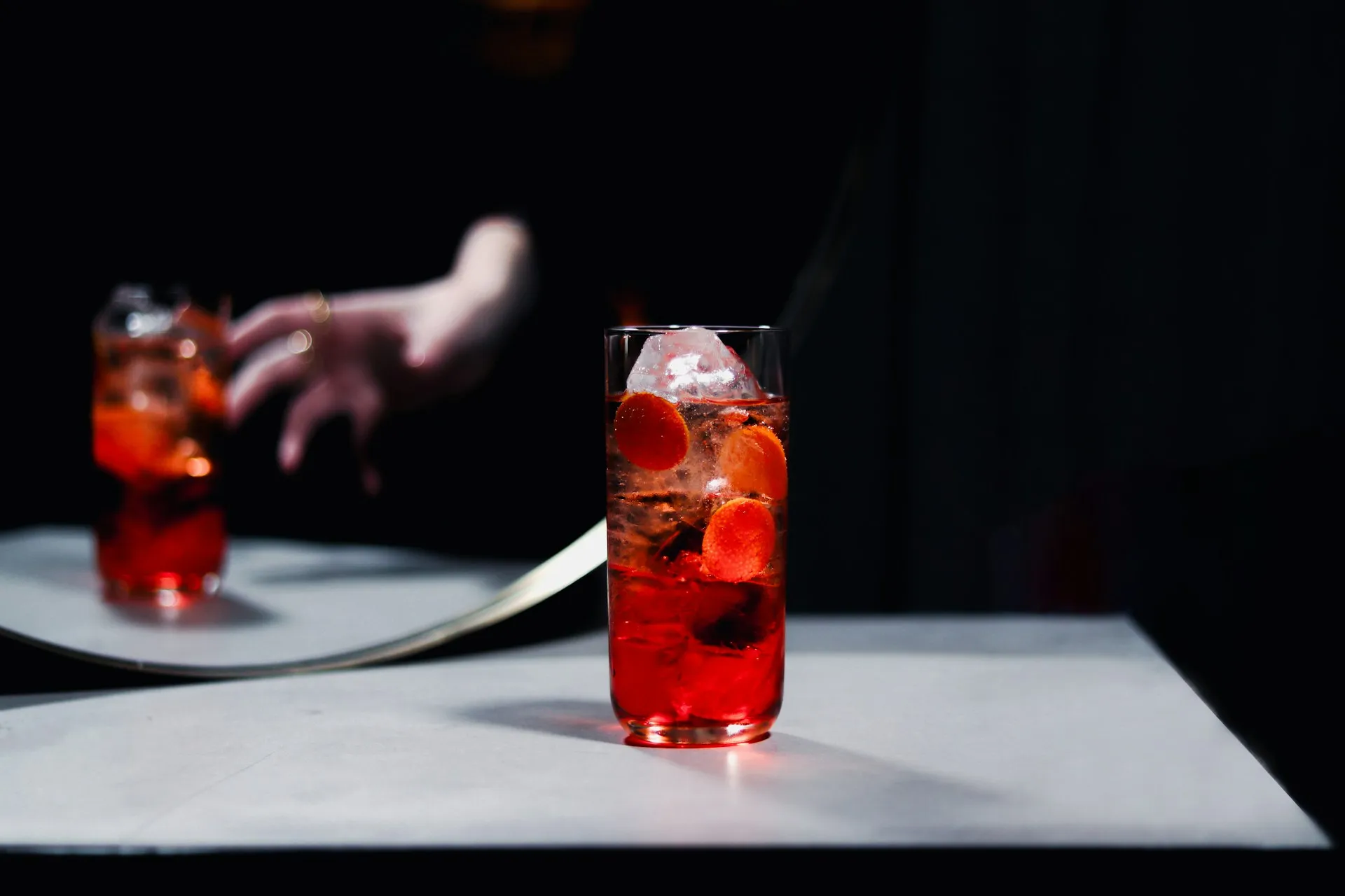 Tall red cocktail with ice and orange slices on a bar counter, with a second drink blurred in the background in a dark lounge setting.