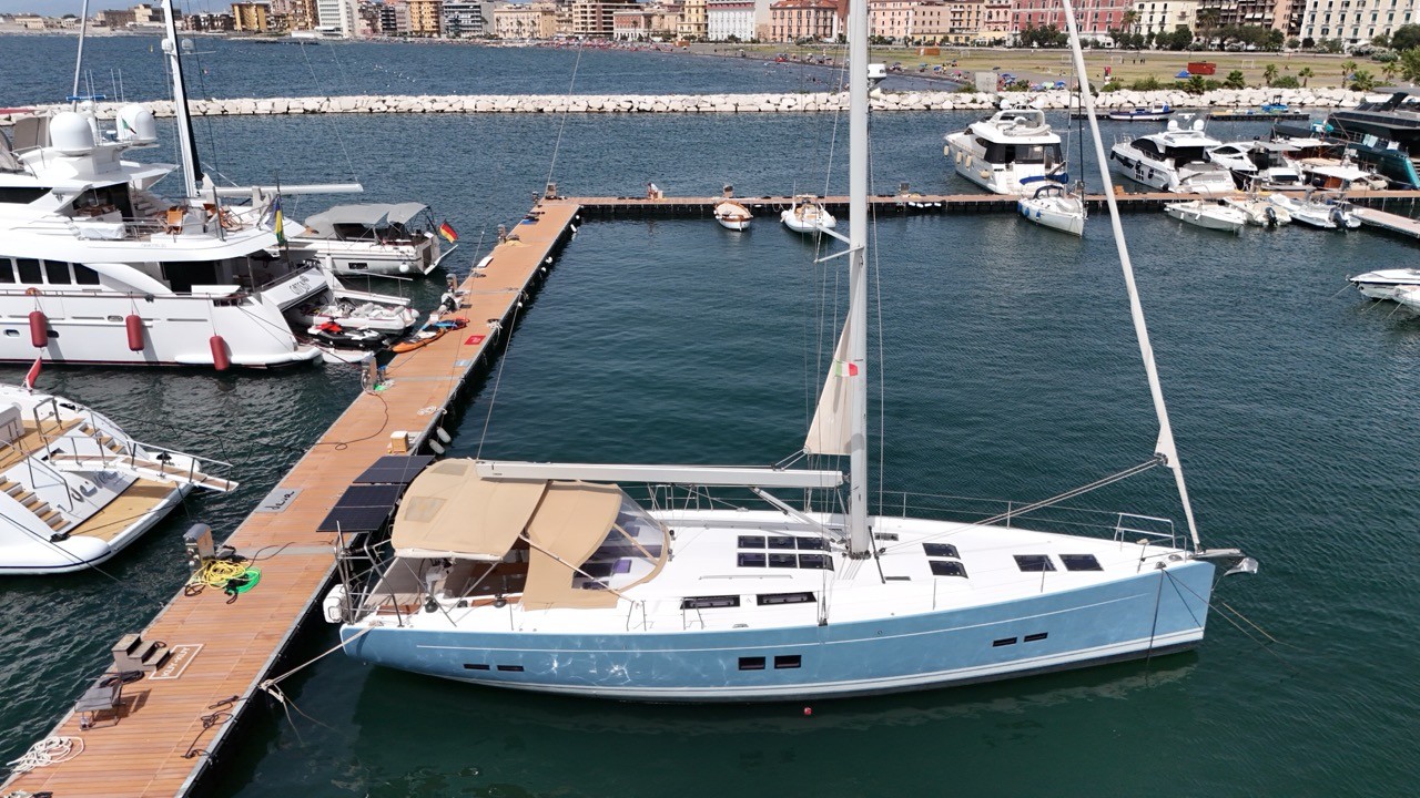 A Hanse 575 sailing yacht with a light blue hull moored bow-to at a teak finger dock in a marina, with superyachts and a city skyline with beachfront buildings in the background.