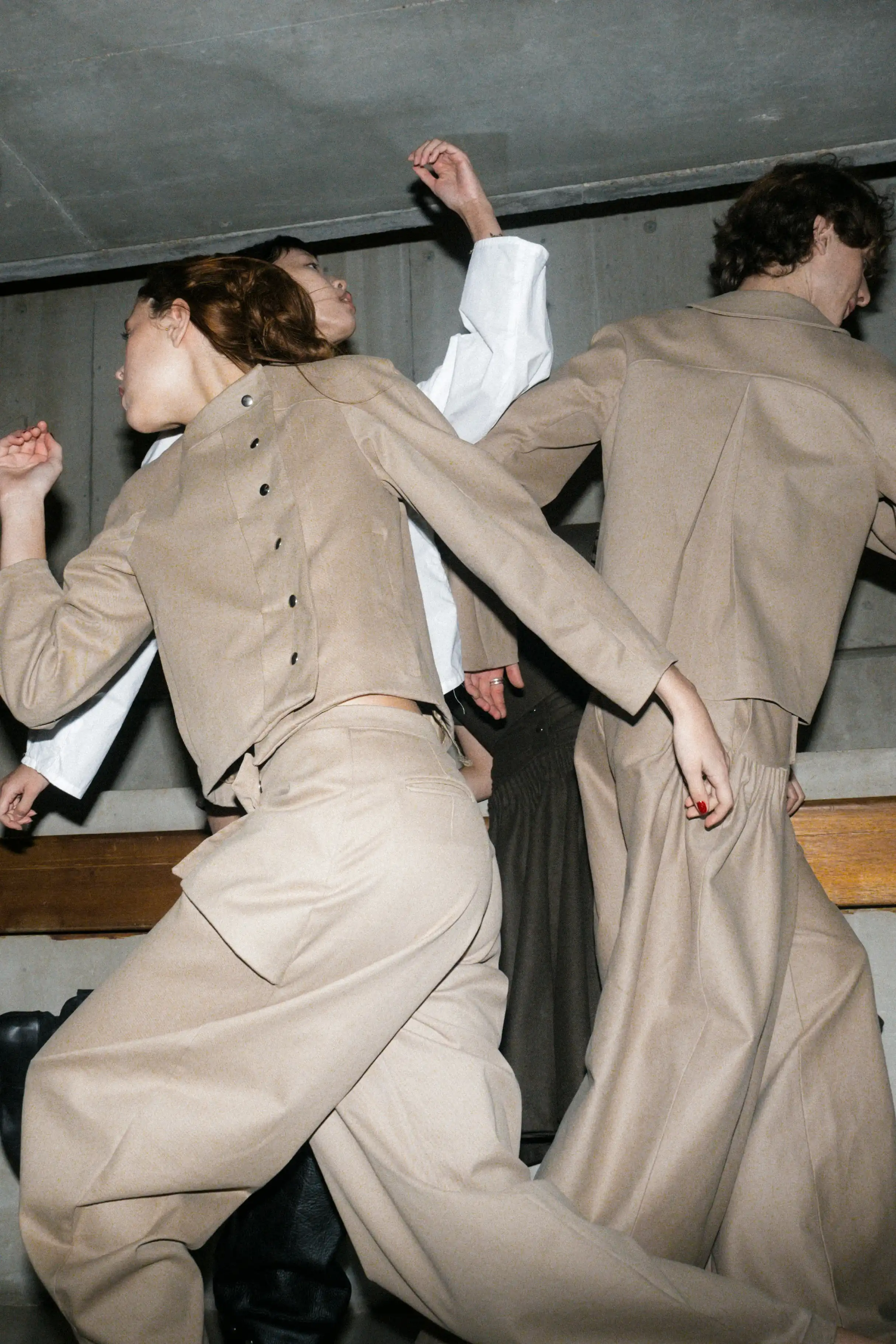 Models in motion wearing matching beige tailored jackets and wide-leg trousers in an industrial setting.