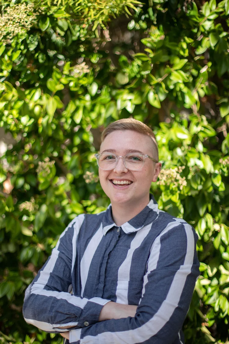 Uma pessoa com cabelo curto e óculos, vestindo uma camisa listrada azul e branca, sorri de braços cruzados diante de uma exuberante folhagem verde.