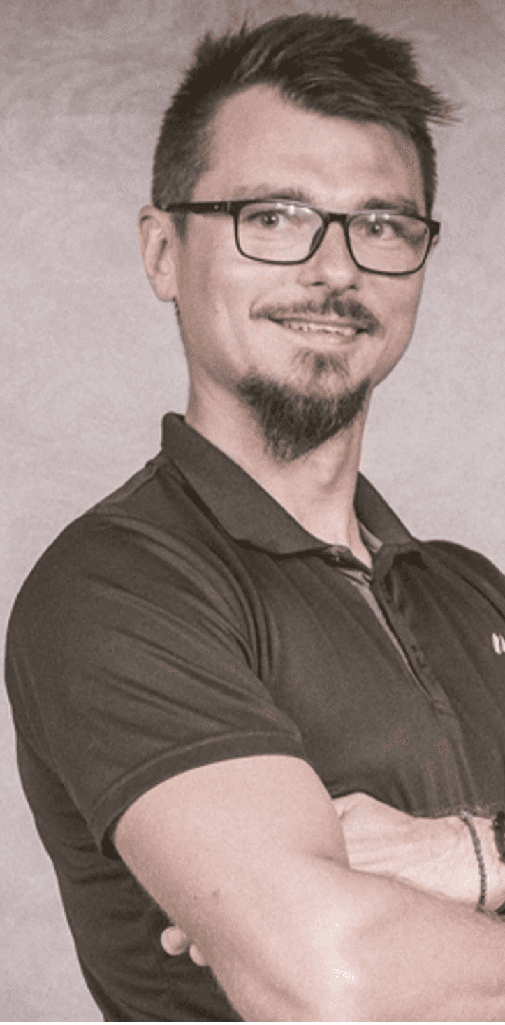 Zdjęcie przedstawia kobietę o jasnych, falowanych włosach i okularach w ciemnych oprawkach. Ma na sobie czarną bluzkę z wzorem i patrzy prosto w obiektyw. Tło jest jednolite, ciemne.