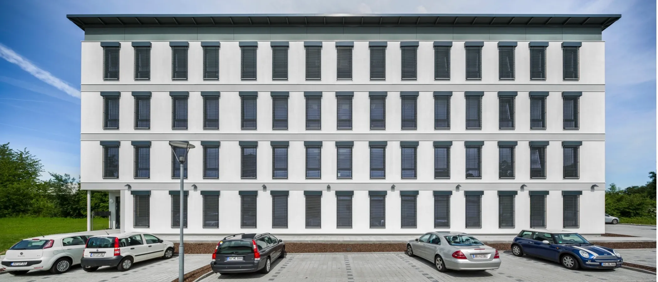 Moderne Fassade eines Gewerbehindos mit mehreren Fenstern, Rollläden und davor parkenden Autos unter blauem Himmel.