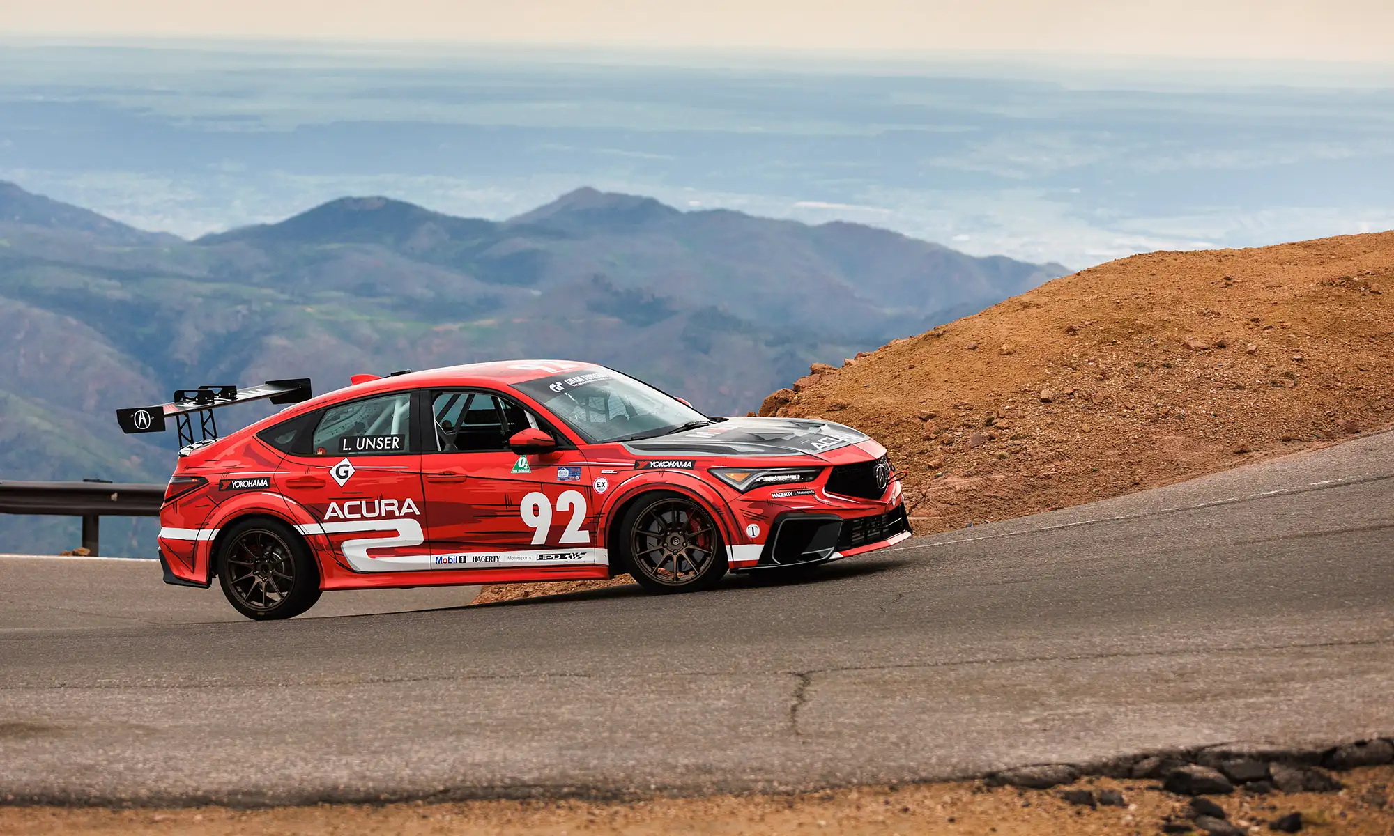 A red Acura race car, driven by Loni Unser, taking a corner at high speed with one wheel lifting off the ground.