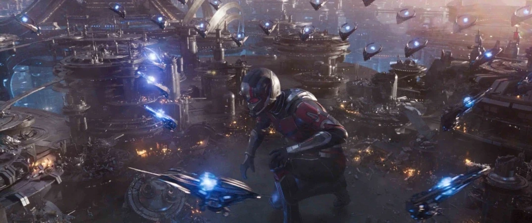 A giant person in a futuristic suit stands among a sprawling sci-fi cityscape under attack. Flying ships with blue lights are visible, and explosions illuminate the scene, creating a sense of high-tech warfare.