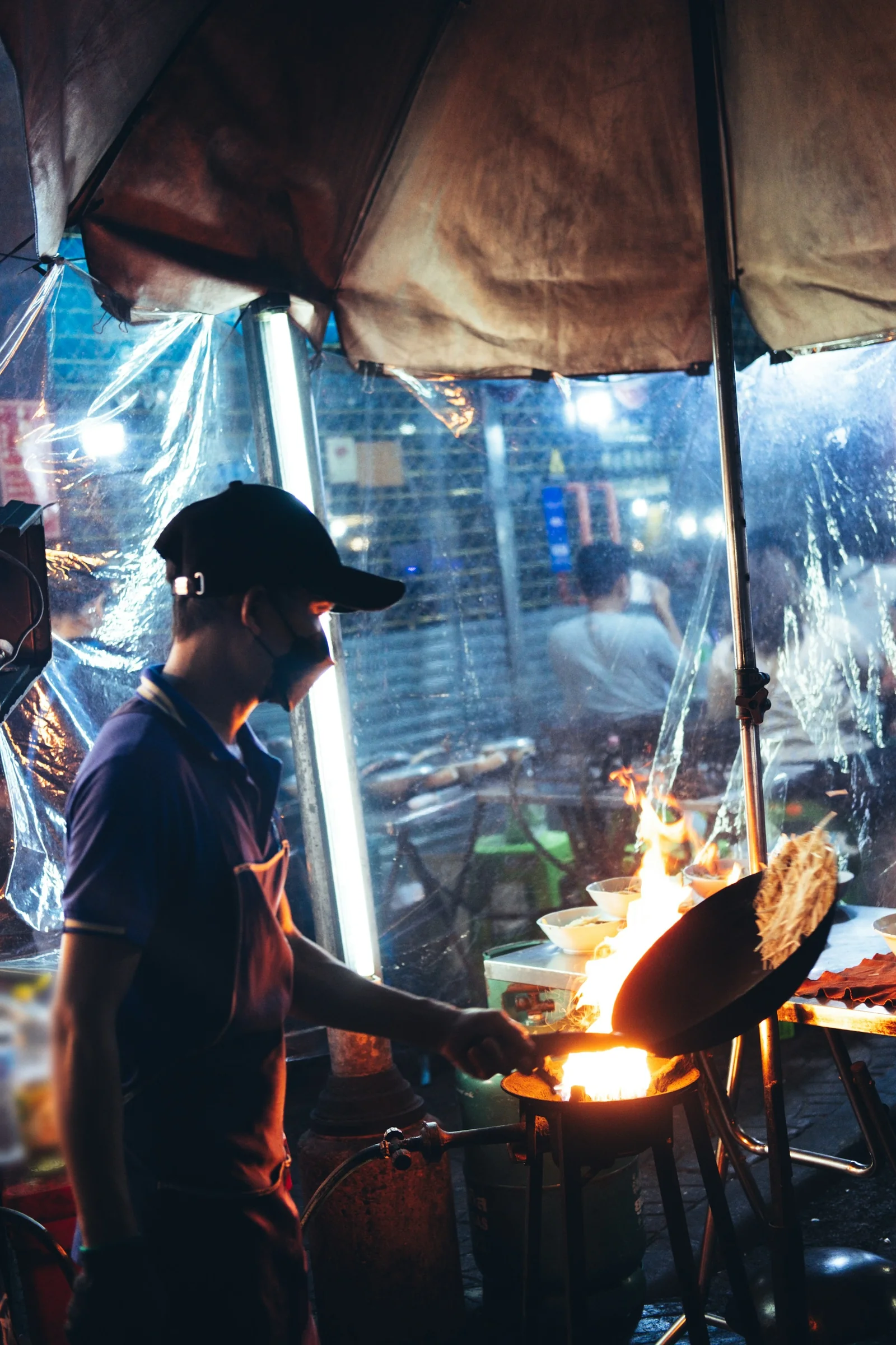 A street cook tossing noodles in a flaming wok at night