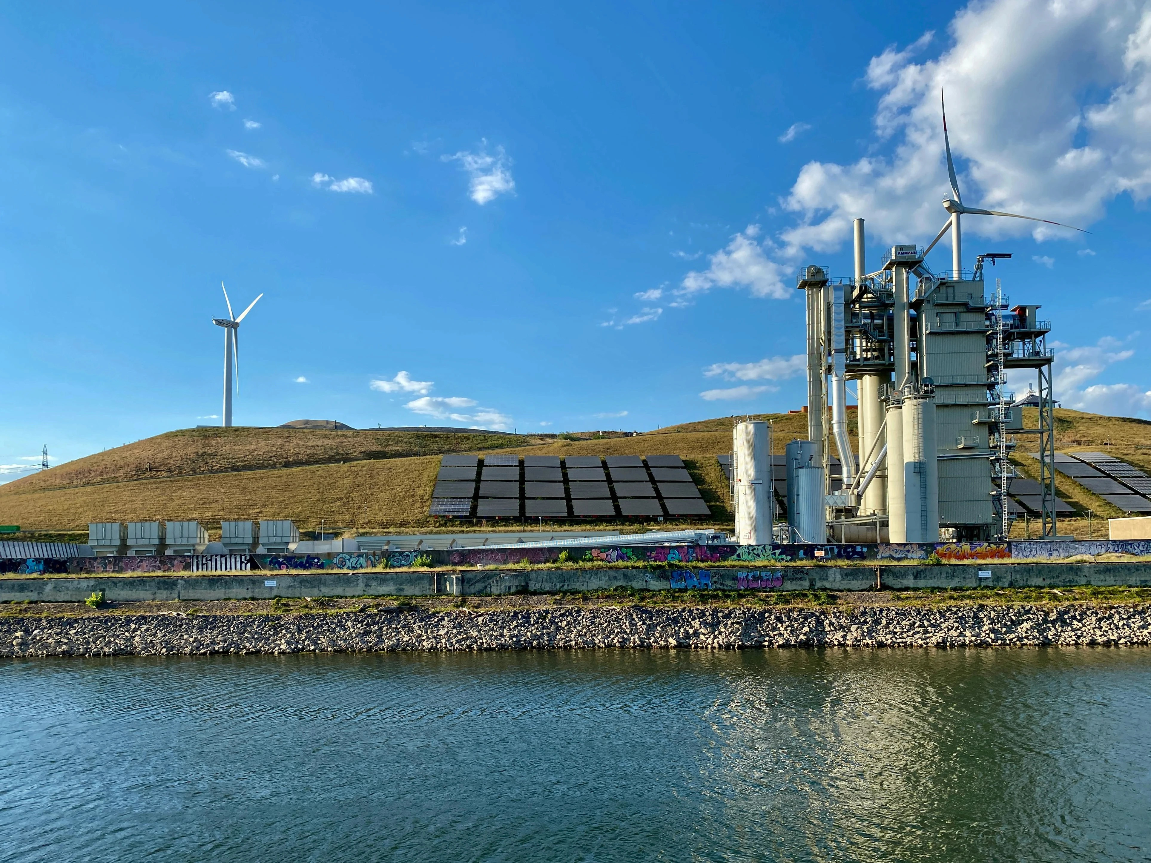 Industrial plant integrated with wind turbines and solar panels on a grassy hillside near a river.