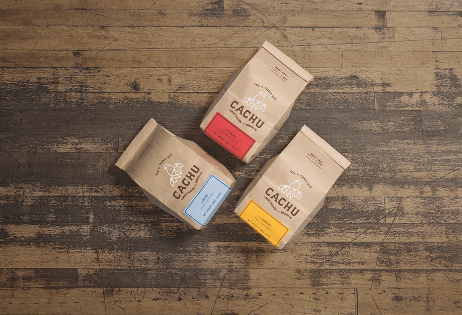 Three brown paper bags labeled “CACHU” are arranged on a wooden surface. The bag on the left is light brown, the middle bag is darker brown, and the bag on the right is orange. Each bag displays a white logo featuring a stylized cactus and text. The bags are slightly angled and appear to be full.