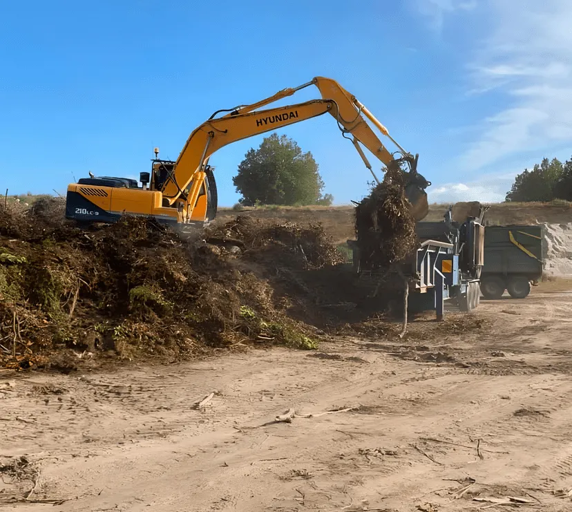 A digger lifting a pile of green waste into a mulcher