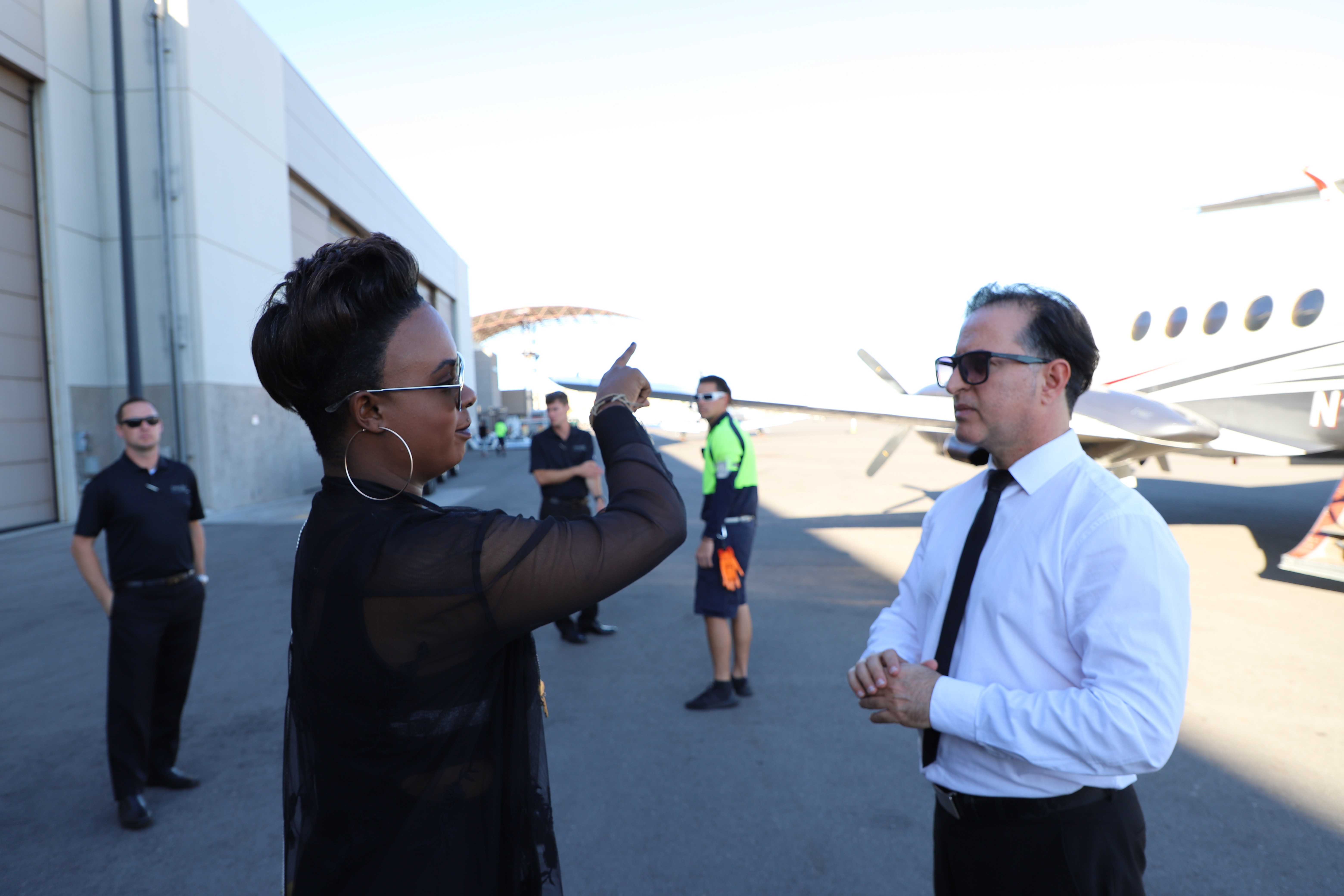 Two individuals are talking outdoors near an aircraft, with others in the background. Bright, clear skies.