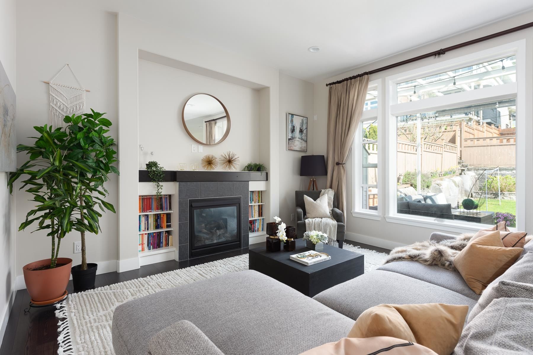 Cozy living room with a gray sofa, a fireplace, large window, plants, and modern decor.