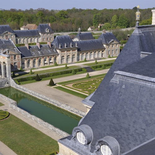 Вид с высоты на величественное поместье с ухоженными садами, геометрически расположенными кустарниками, водными элементами и классическими зданиями со сланцевыми крышами.