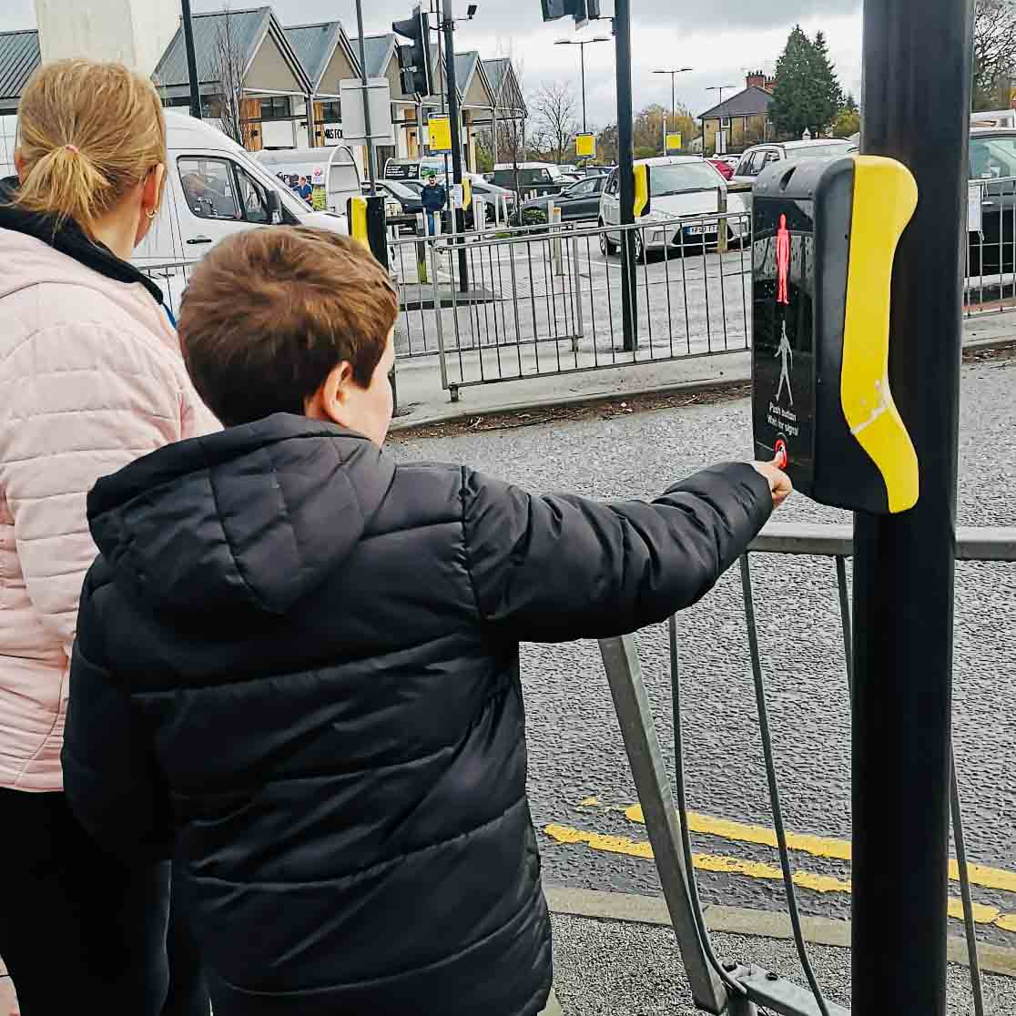 A child is stood at a pedestrian crossing with a Horizon Hub team member, they have pressed the crossing button.