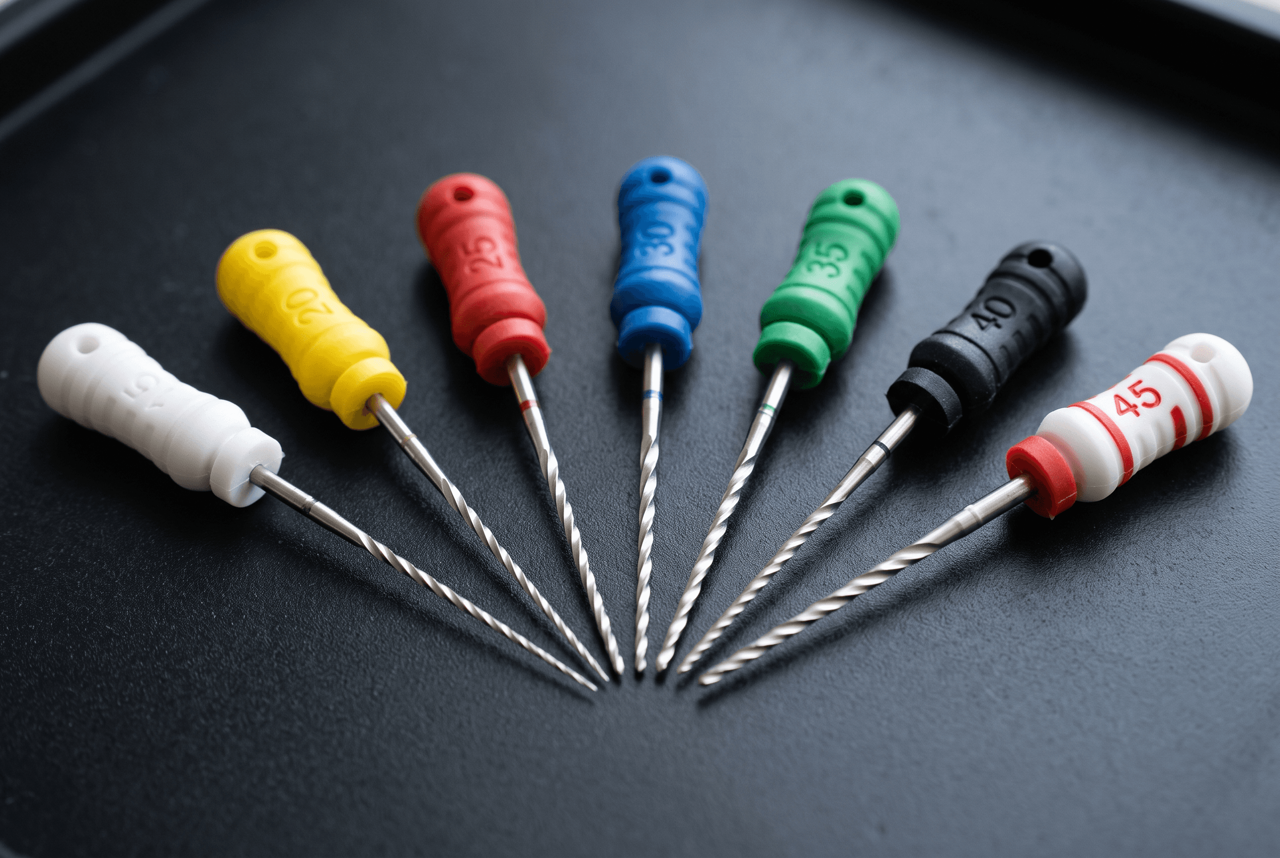 Macro of fine endodontic files fanned out in graded sizes on a dark matte surface, colored silicone handles, precise soft lighting
