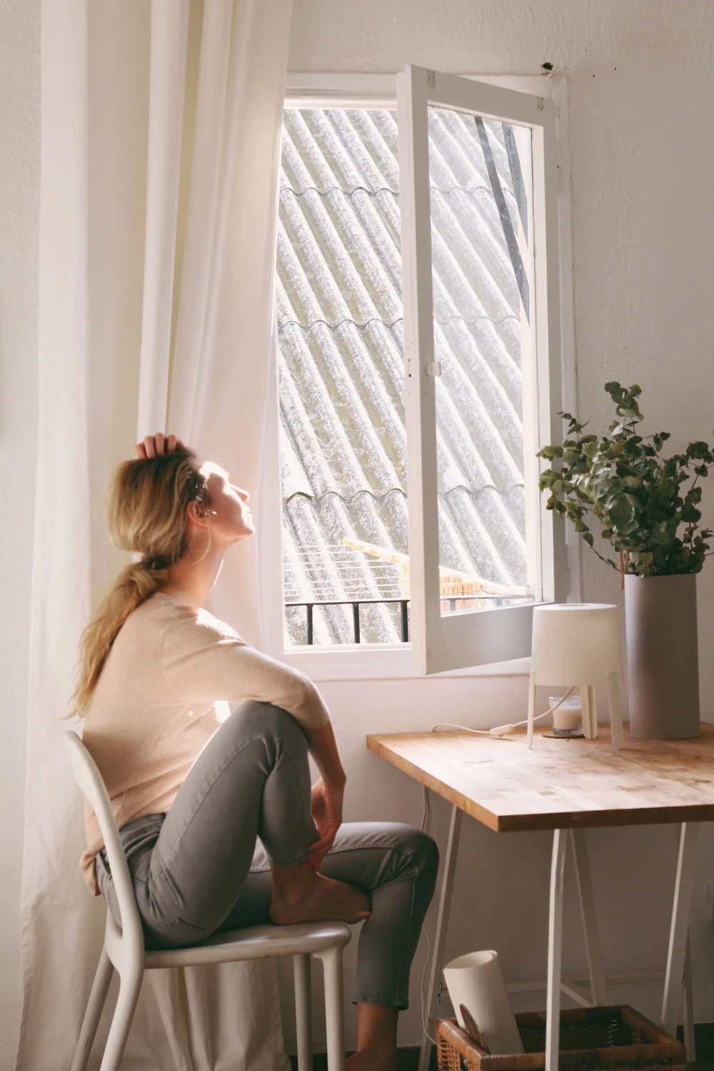 Woman sitting by a bright window, looking outside in a quiet, reflective moment.