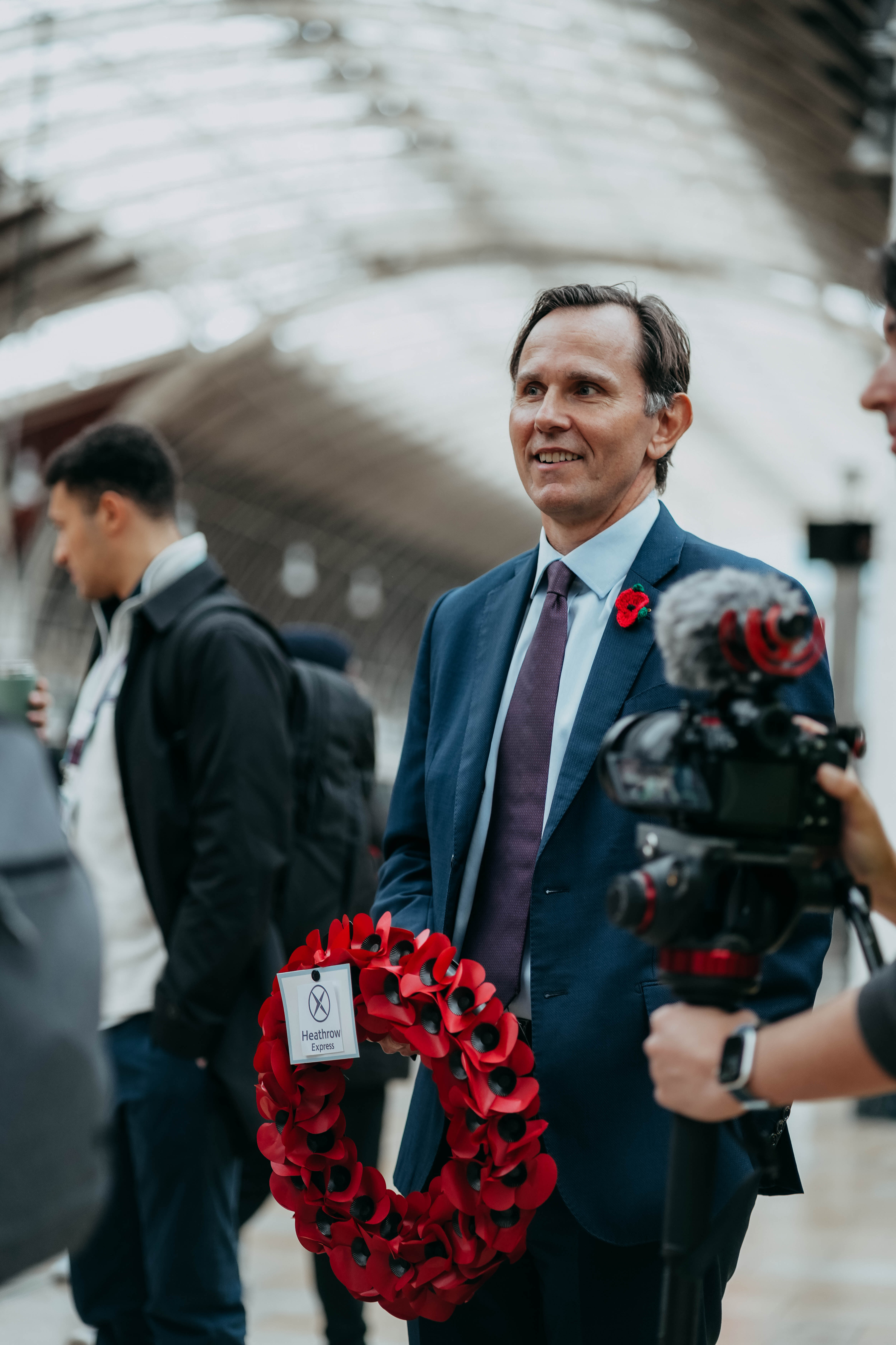 A man in a blue suit with a poppy on his lapel holds a poppy wreath and smiles while being filmed or interviewed in a train station.