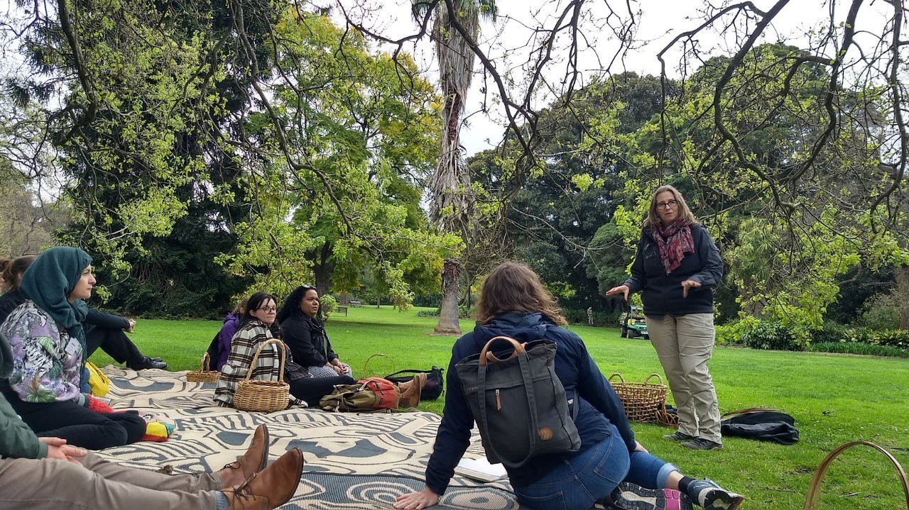 Participantes sentados em toalhas no gramado de jardim botânico ouvem mulher em pé, em atividade ao ar livre