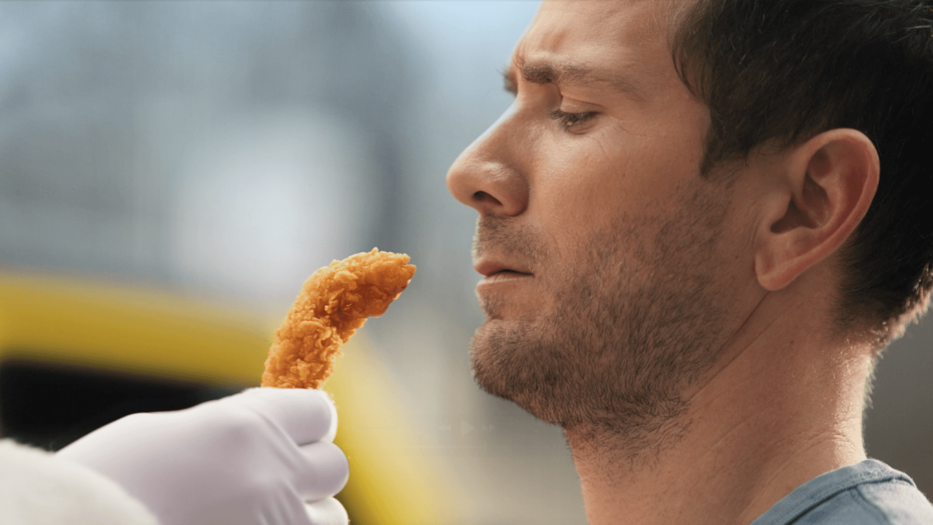 A man stands with eyes closed as a hand offers him a piece of food, set against an outdoor backdrop.