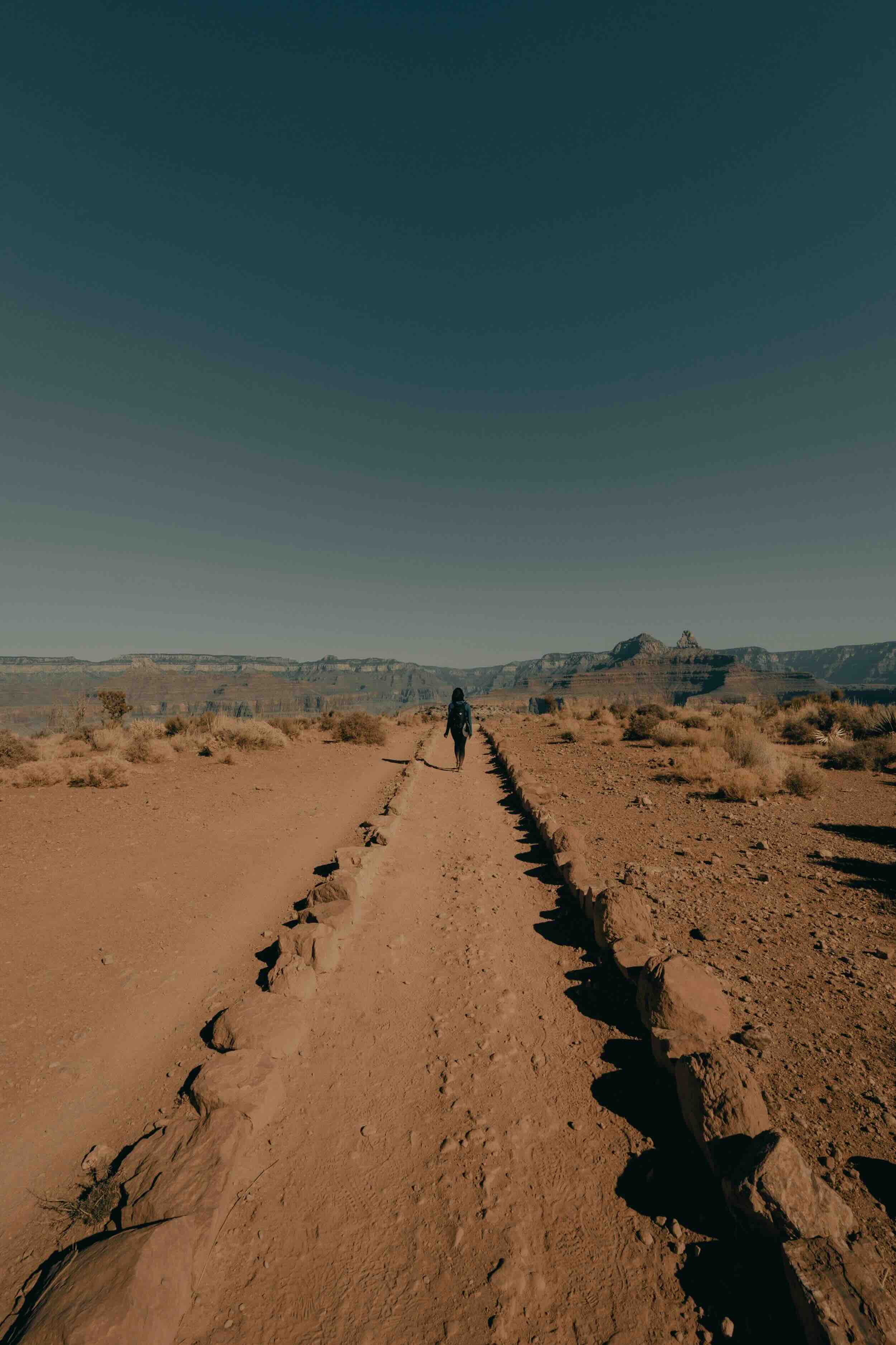 A lone hiker walks along a dusty, rocky trail bordered by stones in a vast desert landscape with rugged mountains in the distance under a clear blue sky