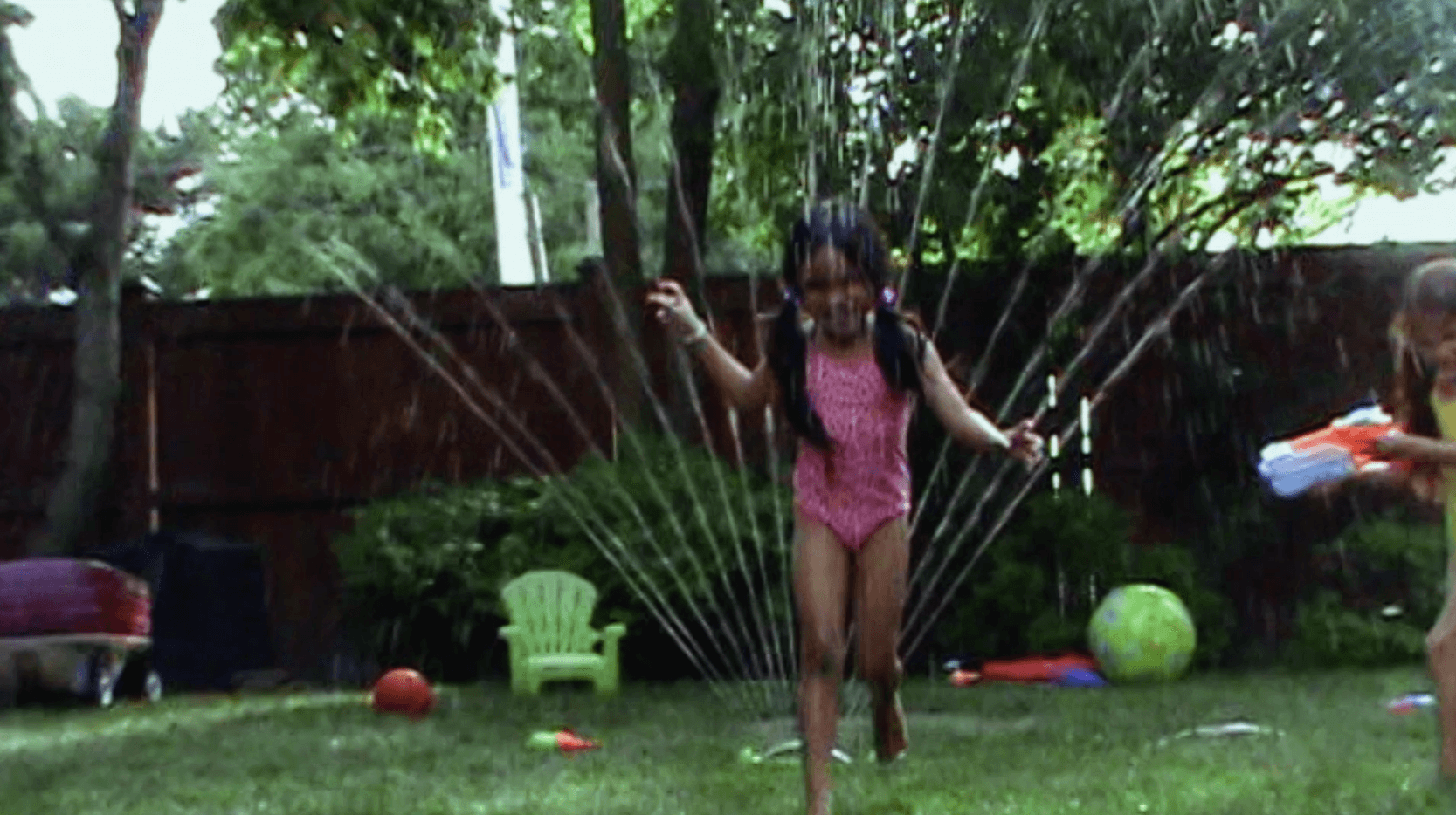 A child in a swimsuit playfully jumps with a water spray in a backyard, surrounded by toys and greenery.