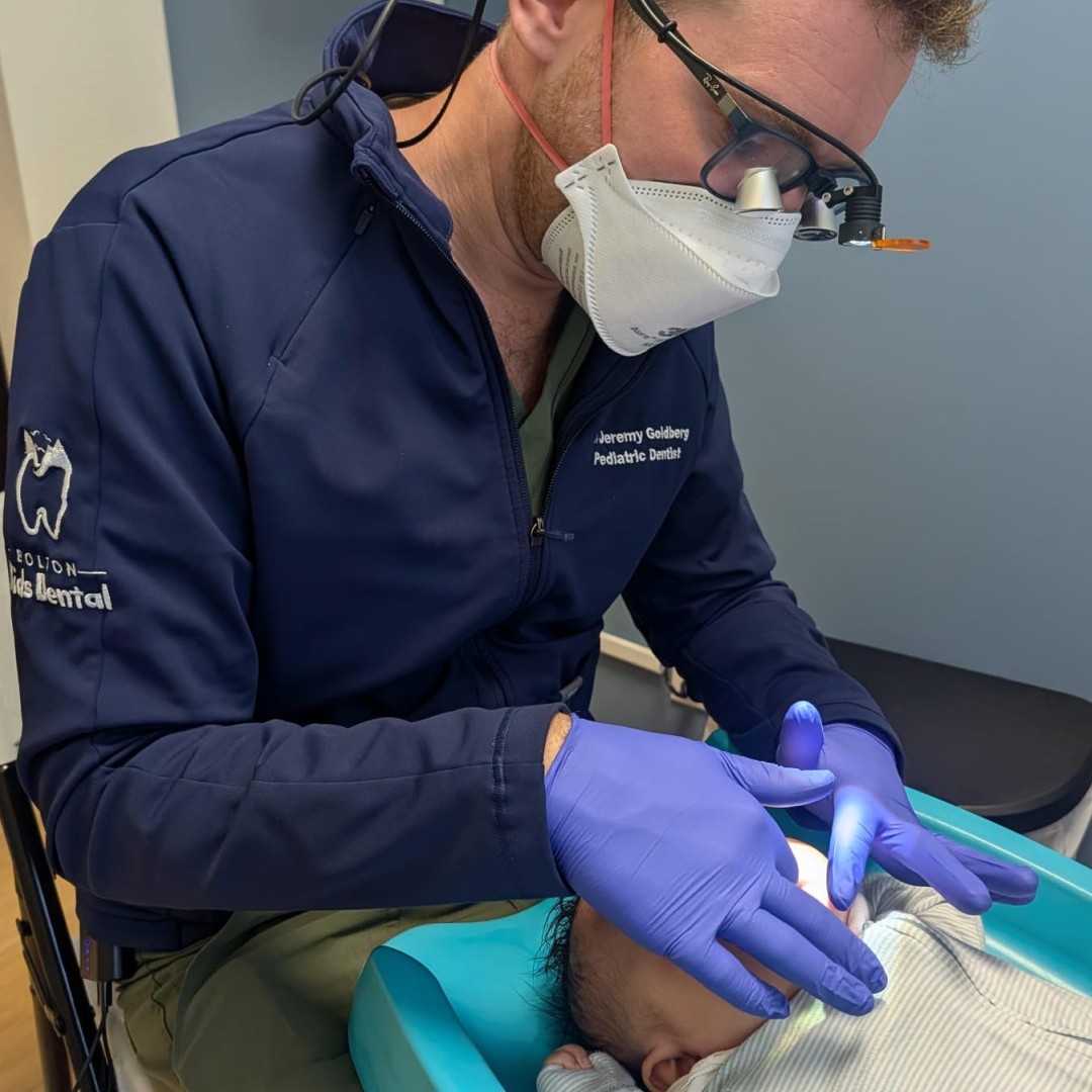 Pediatric dentist performing an infant feeding assessment on a baby in a clinical exam room, wearing gloves, mask, and magnifying loupes.