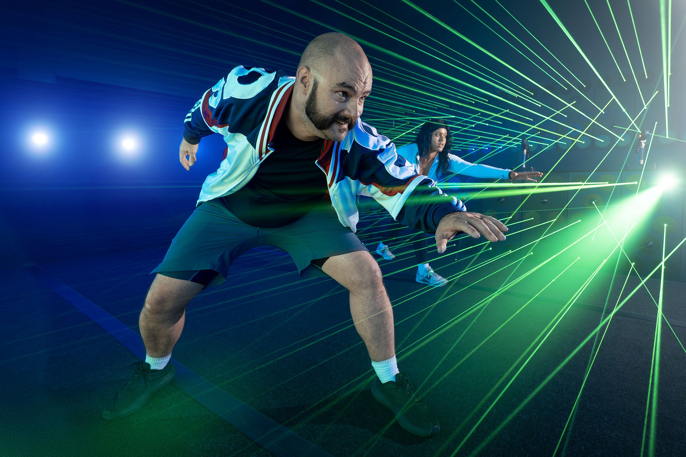 Two people dance and playfully avoid green laser beams in a dark room with bright blue accents.