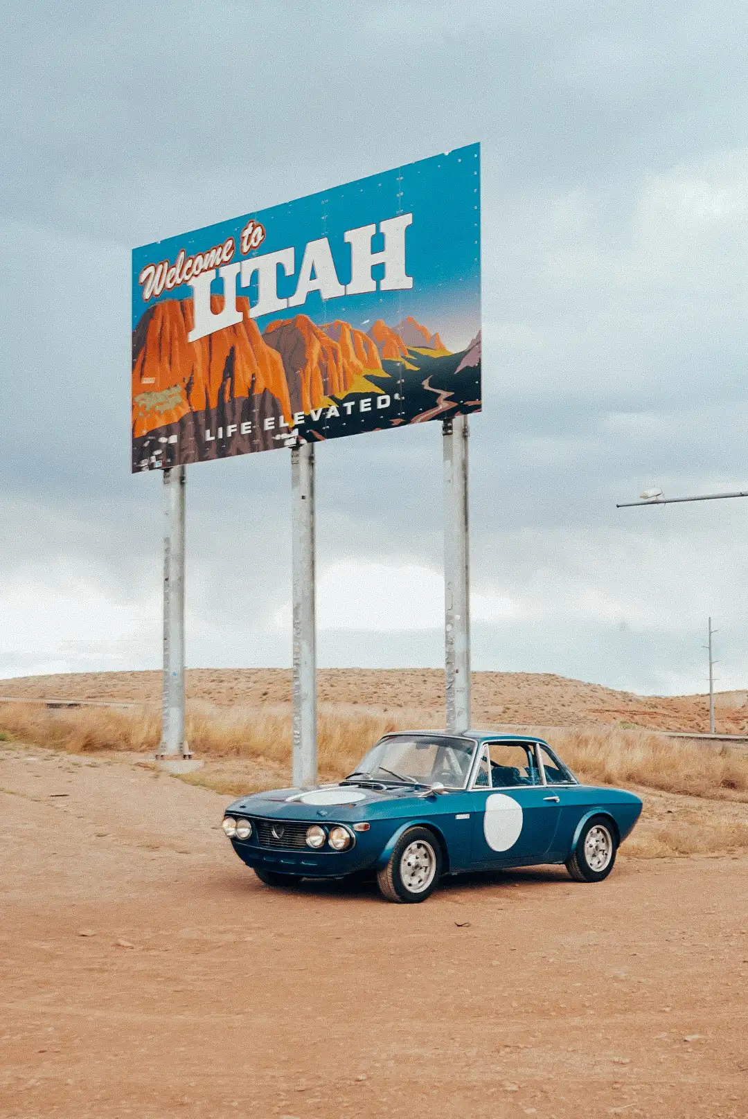 A blue Lancia Fulvia sports car parked under a sign reading, “Welcome to Utah.”