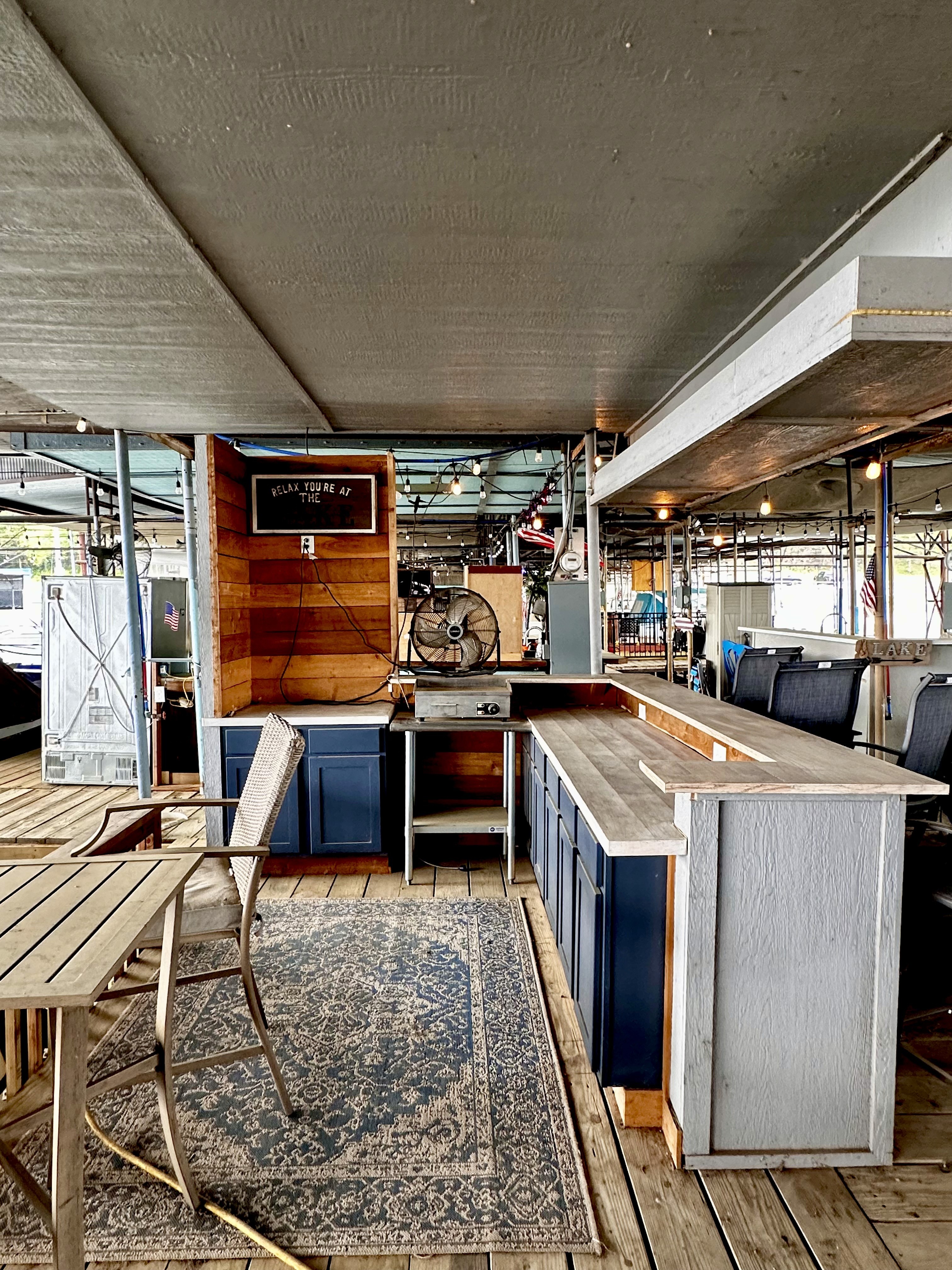 Rustic outdoor bar area under a covered wooden deck with blue cabinets, a wooden bar top, a ceiling fan, and a cozy seating area featuring a wooden table and chairs, set against a background of string lights.
