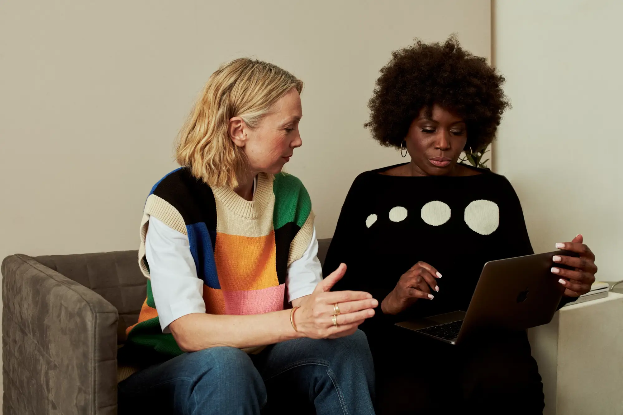 two woman sitting on a couch looking at a laptop smiling in conversation