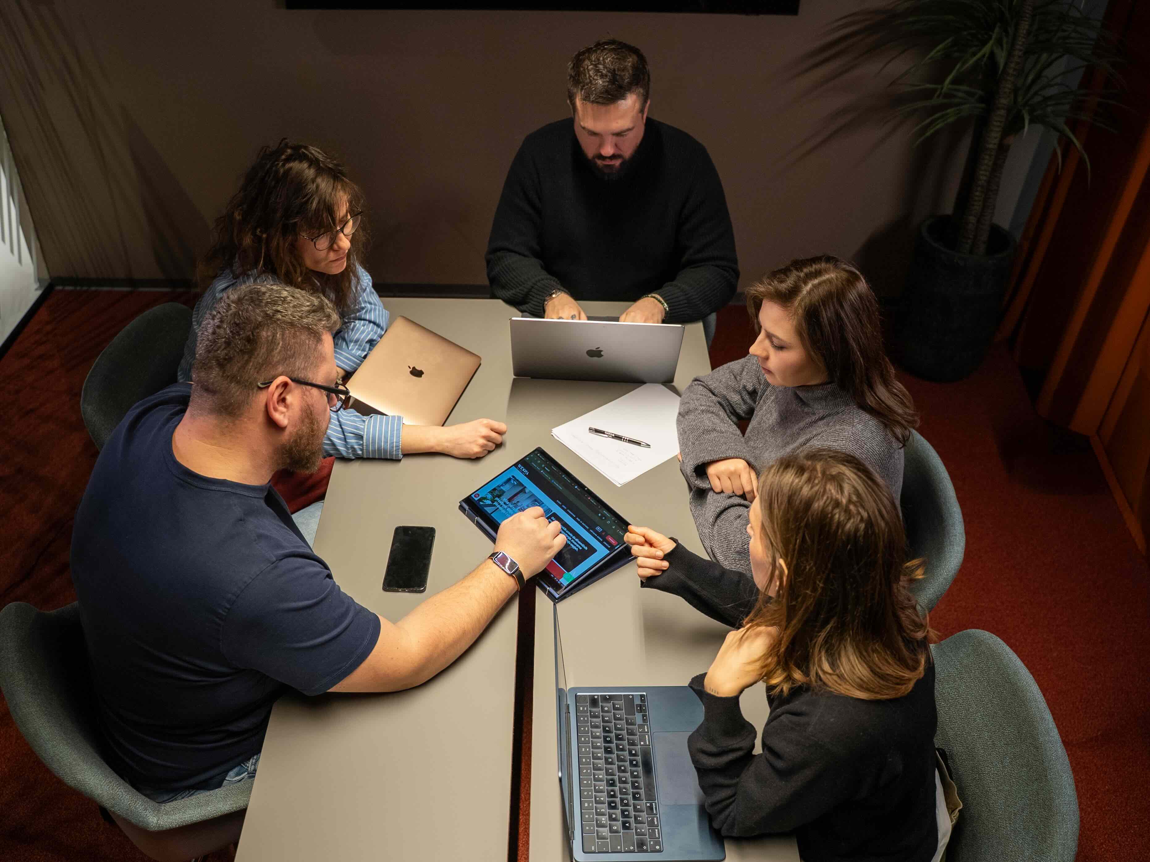 Young man in a green hoodie holding a yellow phone while seated at a table with a tablet.