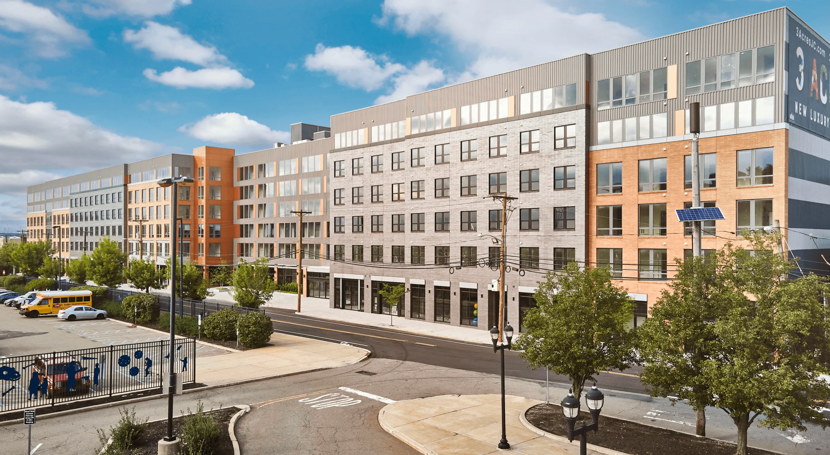 Modern multi-story building surrounded by trees and a clear blue sky. Street visible in the foreground.