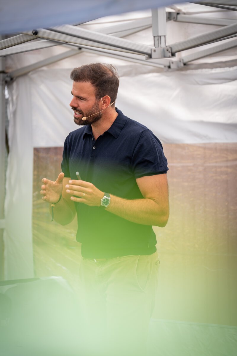 Max Planer speaking at an outdoor event, wearing a headset microphone