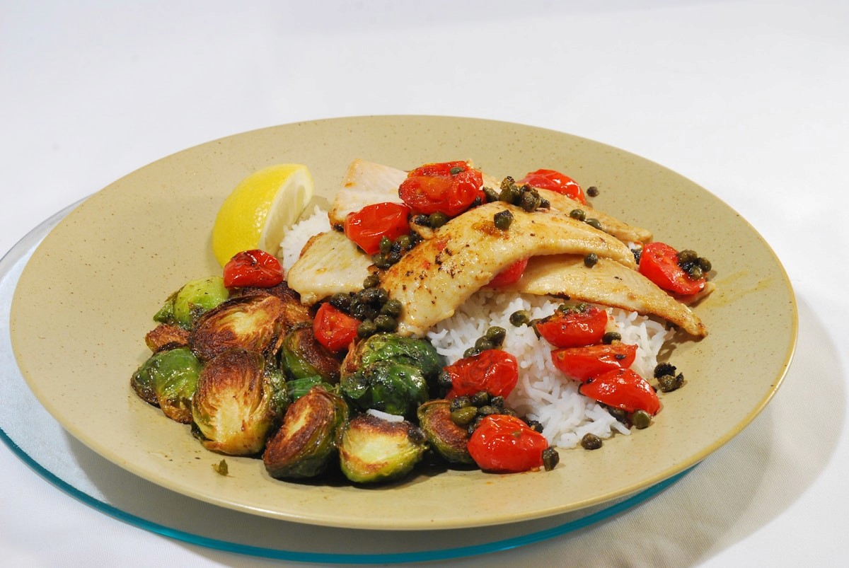 A balanced plate of cooked fish over rice with roasted brussels sprouts and tomatoes, showing a well-rounded meal of lean protein, whole grains and vegetables.