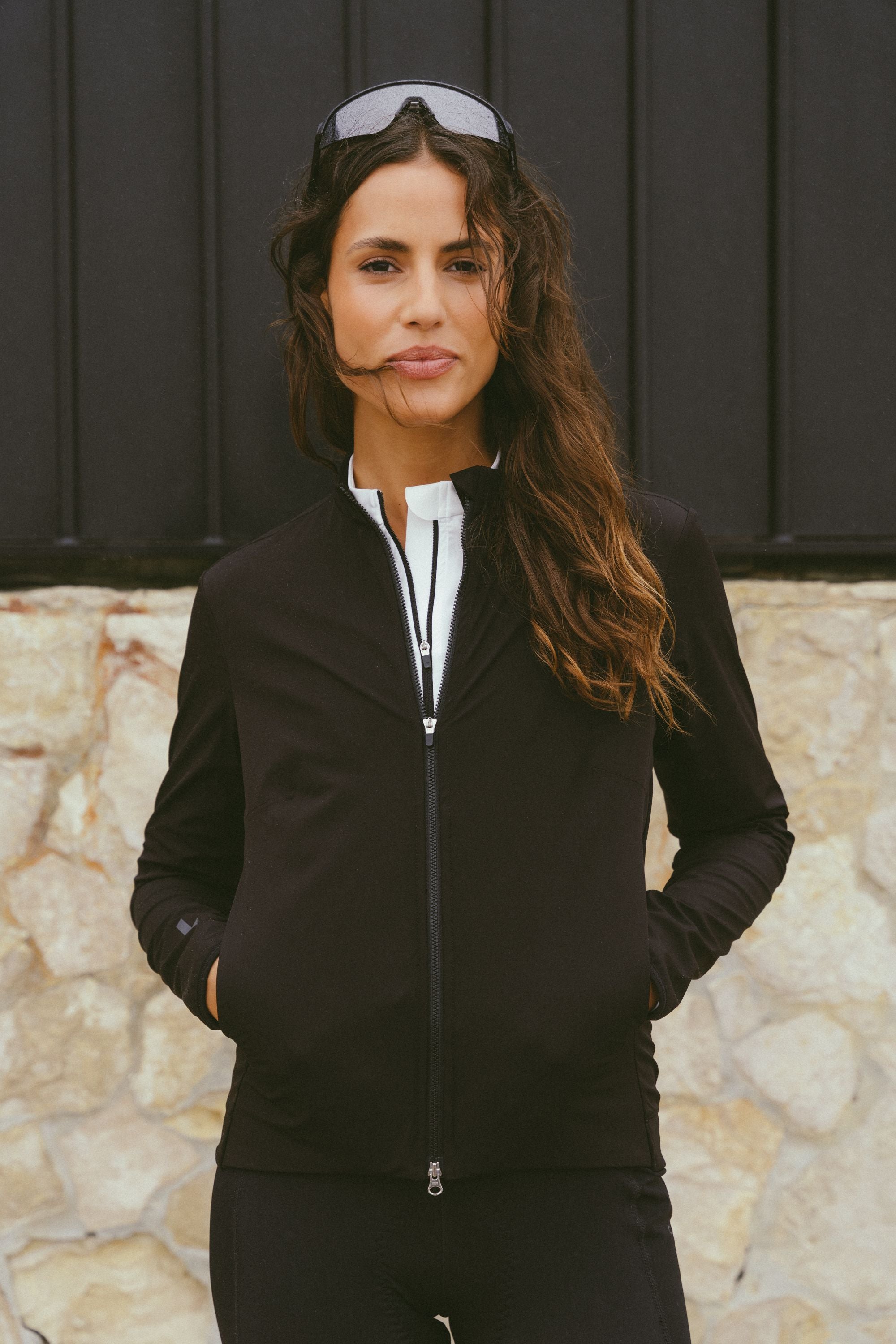 Front-view-of-a-confident-women-wearing-the-Black-Momentum-jacket-zipped-halfway-with-her-hands-in-the-pockets-a-white-shirt-undermeath-and-goggles-pushed-up-onto-her-head