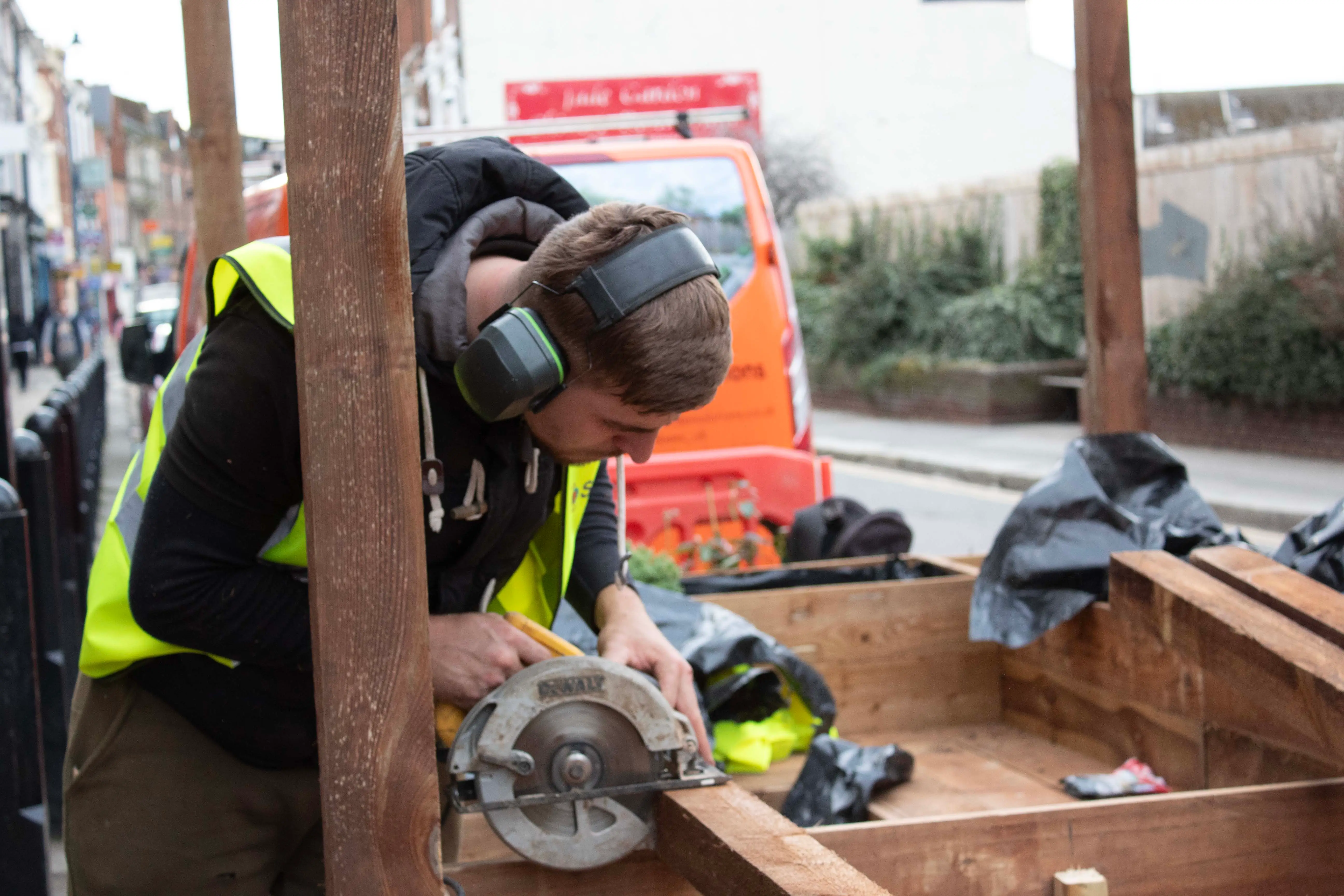A construction worker in a safety vest is kneeling and using a tool on a wooden structure at a work site.