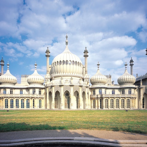 A large, ornate building with domes and intricate architecture set against a partly cloudy sky, with a grassy area in front.
