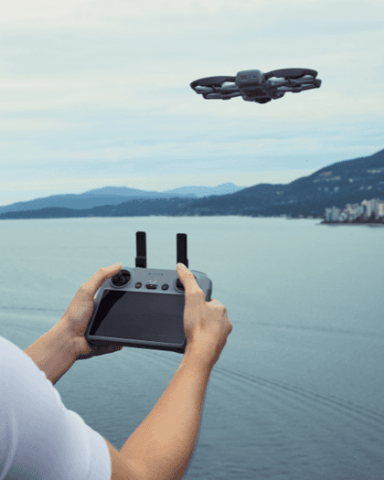 A person holding a drone controller with a drone flying above a calm lake, surrounded by mountains and city skyline in the background.
