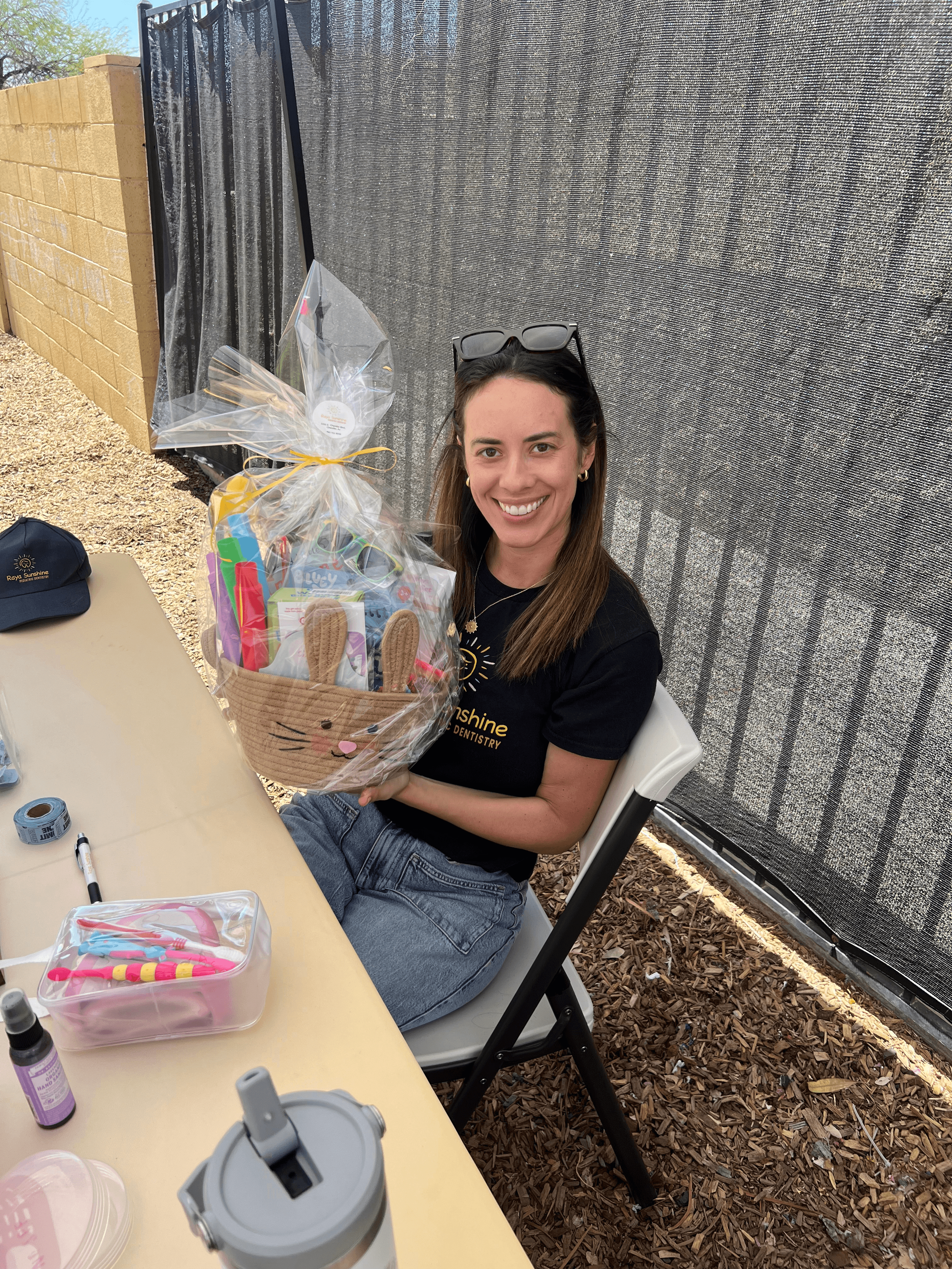 Dr. Raya smiling sitting outdoors next to a table with gift bags and event materials