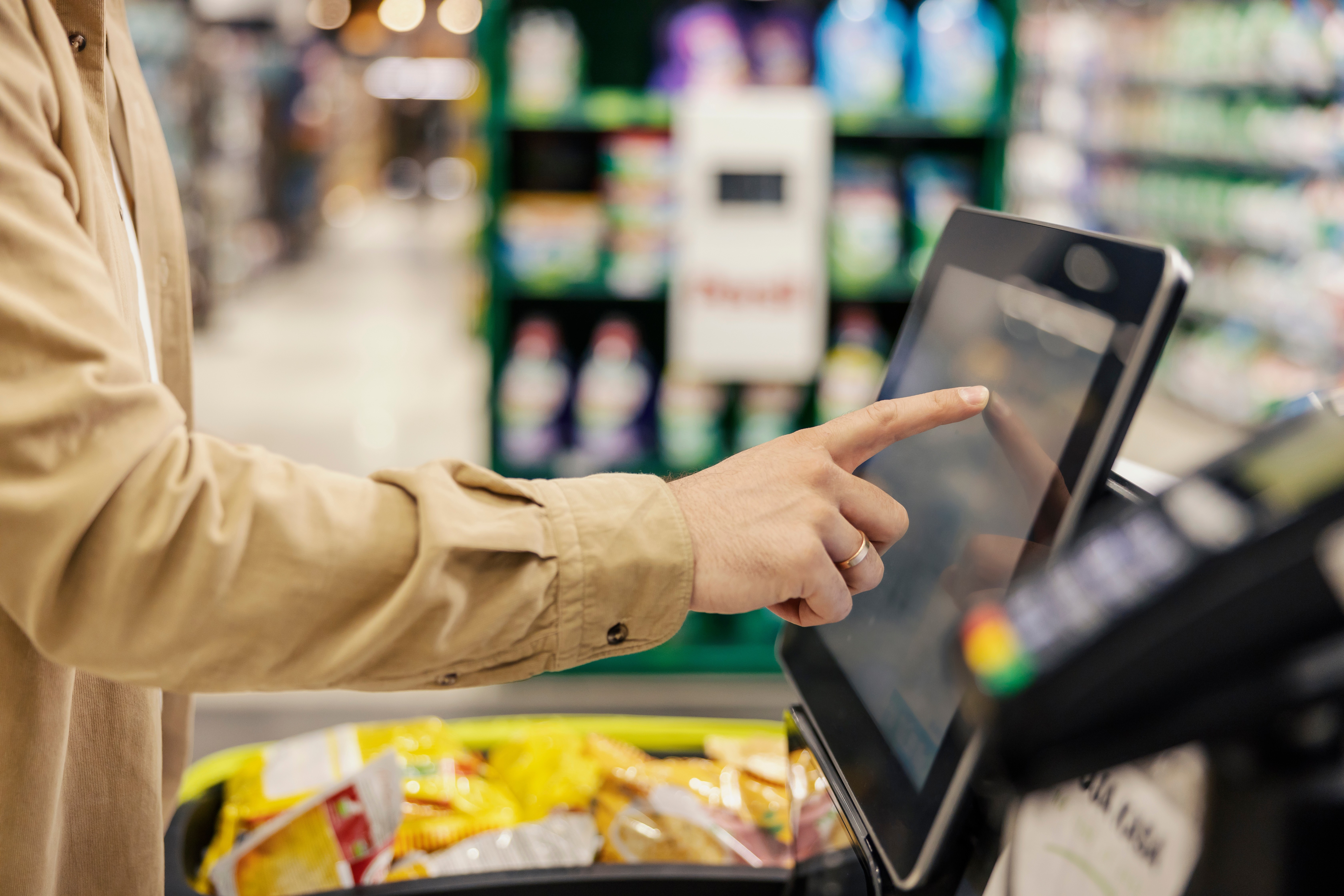 The picture shows a person at a self-checkout kiosk in a retail store. The person is making a selection on the screen.