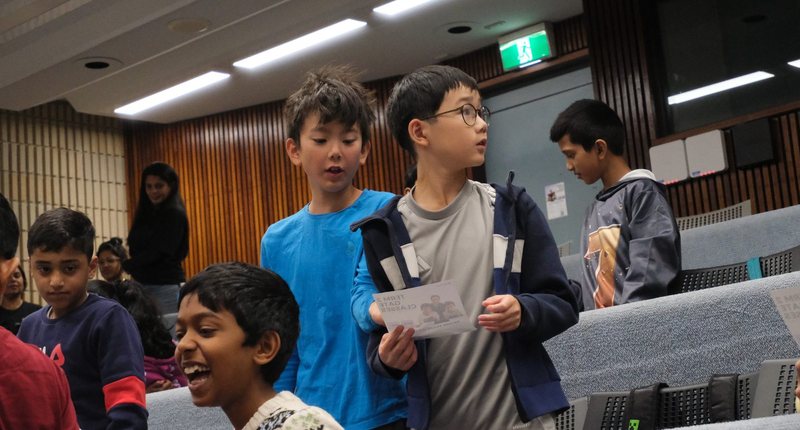 Primary school students standing between the tiered seats of a lecture theatre before an exam, one holding a printed card