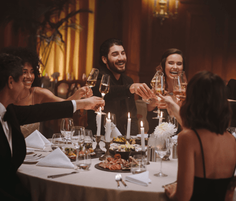 A group of six people are gathered around an elegantly set dining table, smiling and toasting with glasses of champagne. The table is adorned with fine diningware and a variety of dishes. The atmosphere is festive, with soft lighting and a luxurious setting, suggesting a celebration or special occasion.