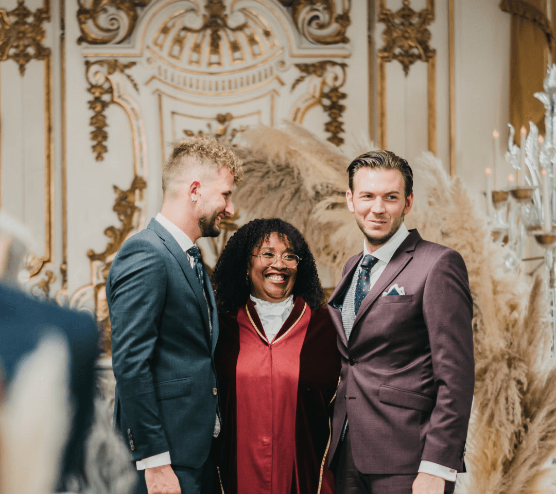 Drie mensen staan samen, gekleed in formele kleding, tegen een elegante achtergrond met decoratieve details het is een stijlvolle gay bruiloft vol liefde en elegantie.