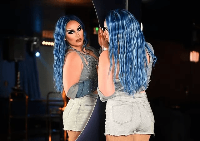 A drag performer with vibrant blue wavy hair and dramatic makeup looking at her reflection in a large mirror, wearing a distressed denim two-piece outfit.