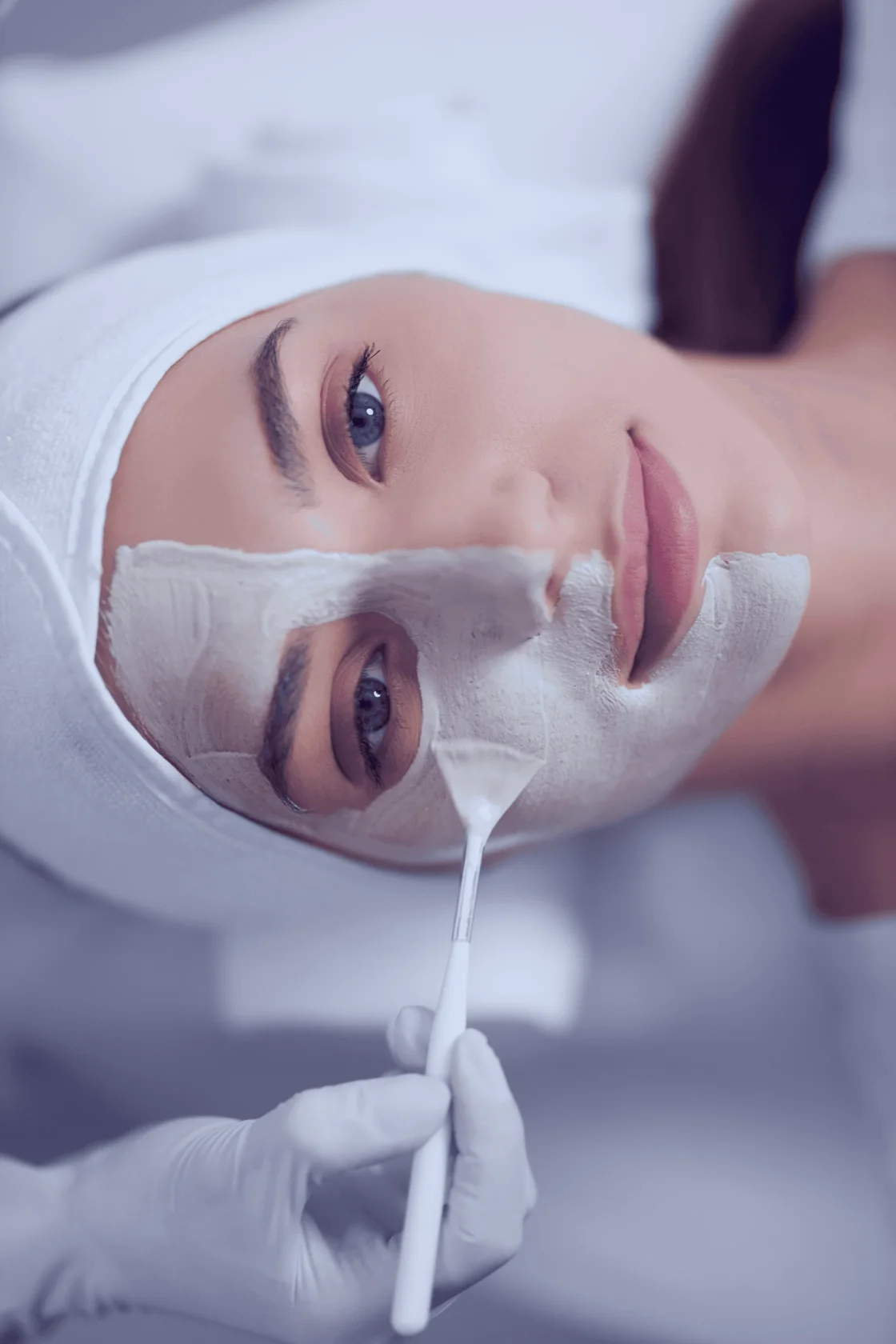 A person receives a facial treatment, lying down with a white headband, as a gloved hand applies a smooth, creamy facial mask with a brush over the person's face.