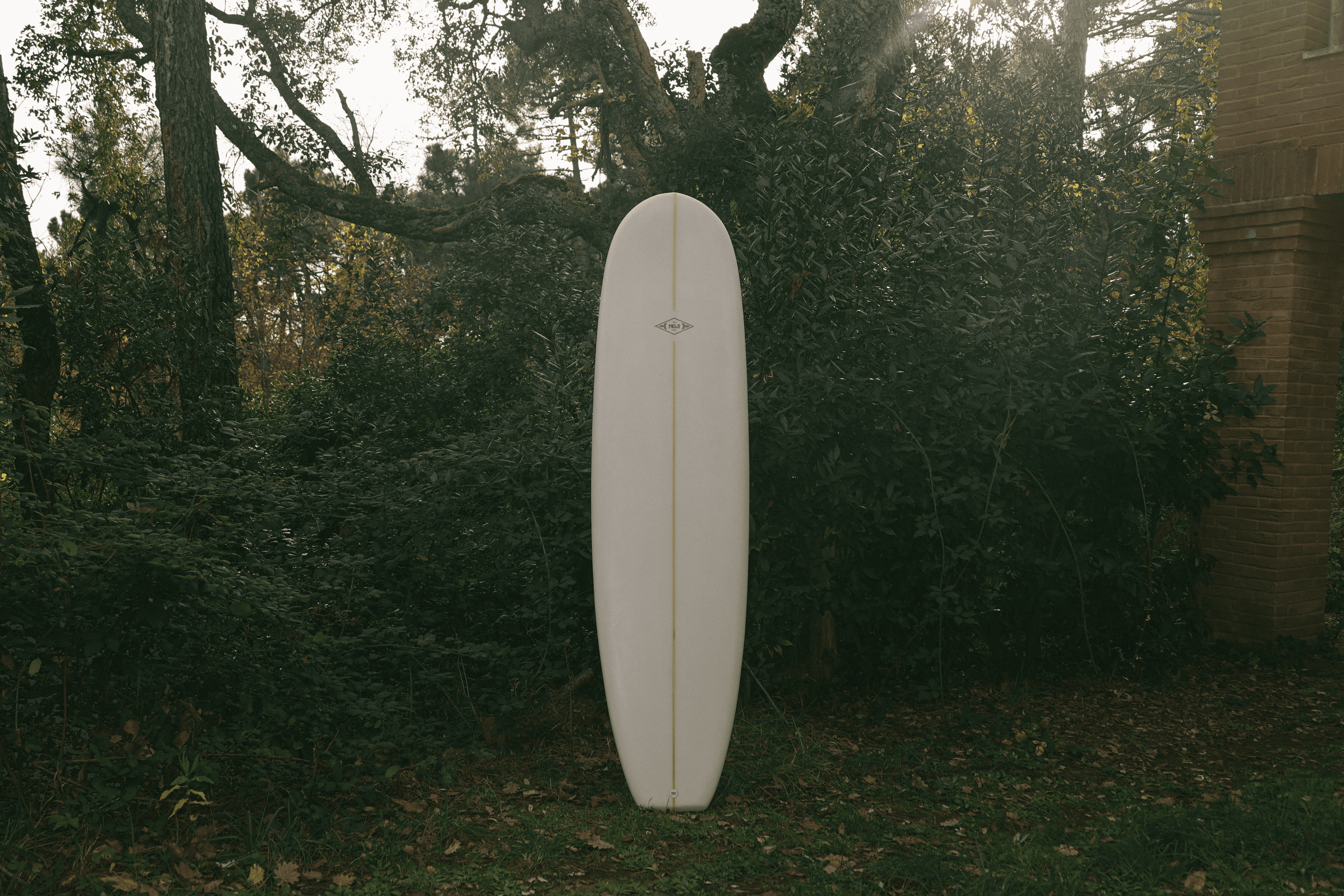Tavola da surf modello Platano bianca con stringer centrale e logo Moji, esposta verticalmente in un giardino boschivo.