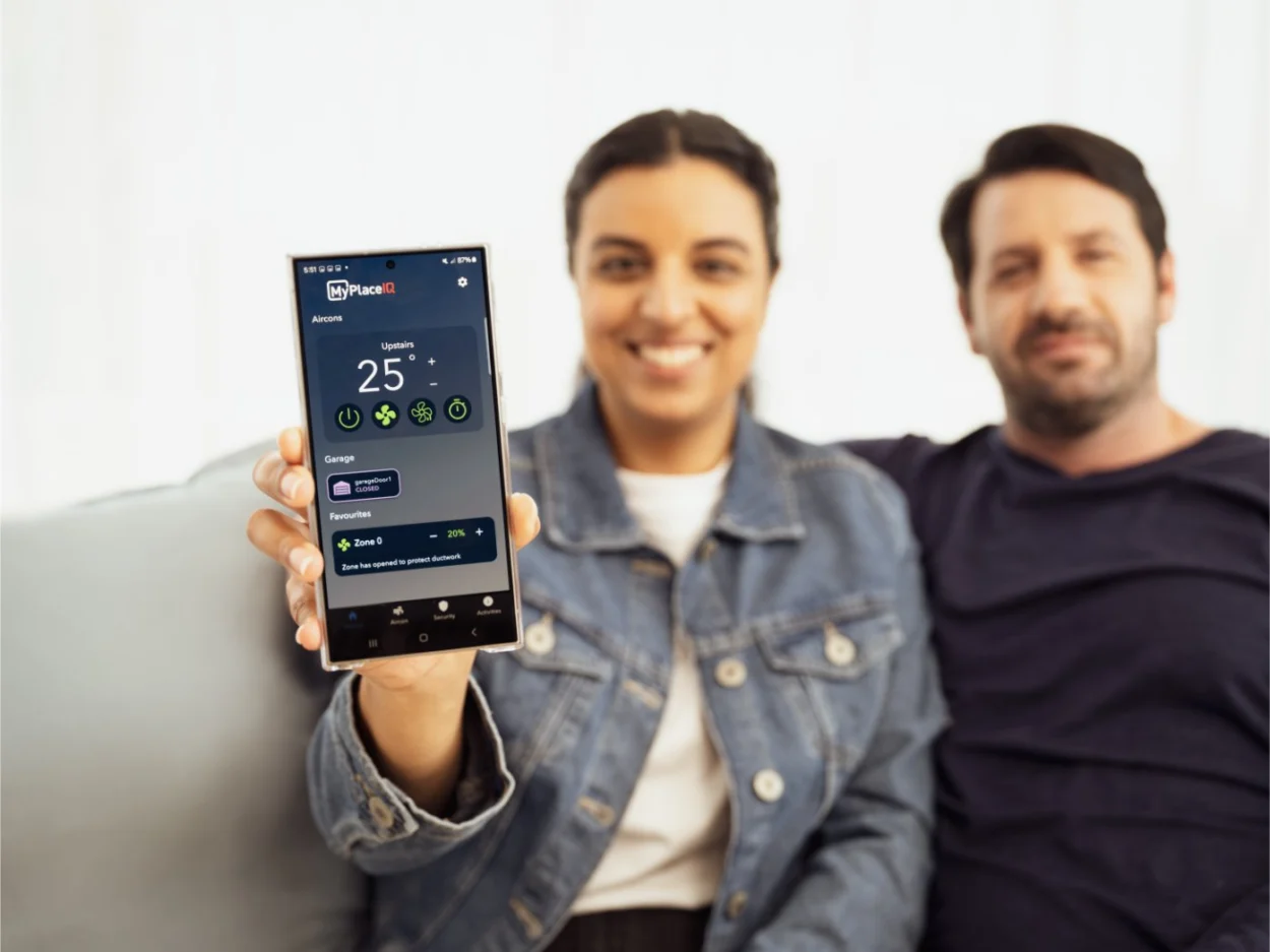 Two people holding up a smartphone displaying the MyPlaceIQ app, illustrating the 2024 launch of MyPlaceIQ, the super smart air-conditioning system with full smart home capability.
