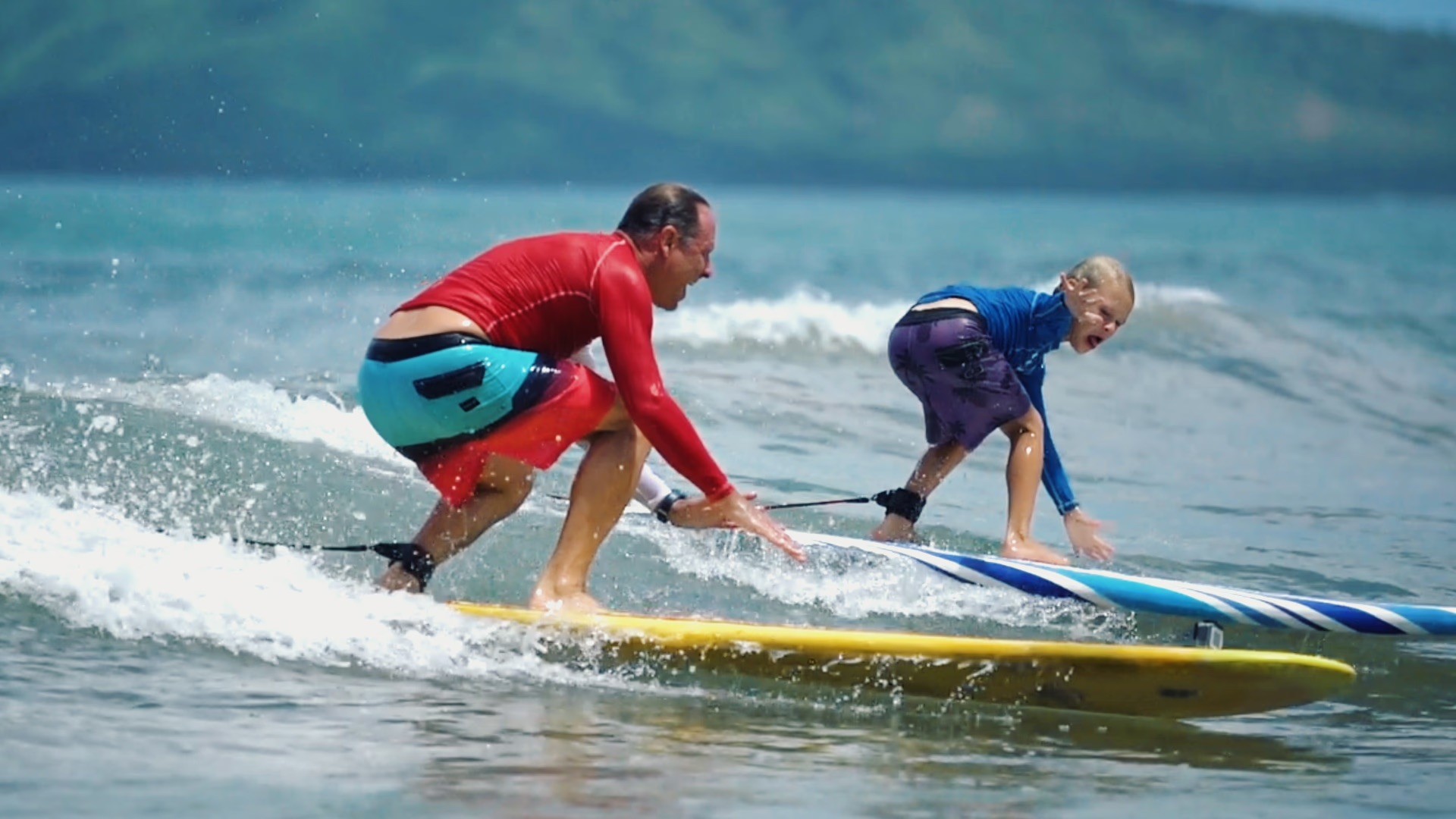 A man and a young boy catch a wave together on separate surfboards in tropical blue water.