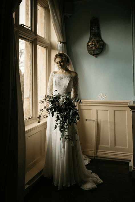 Timeless bridal portrait with a bride in lace, holding a cascading bouquet by a large window, capturing the soft and romantic tones characteristic of vintage wedding photography.